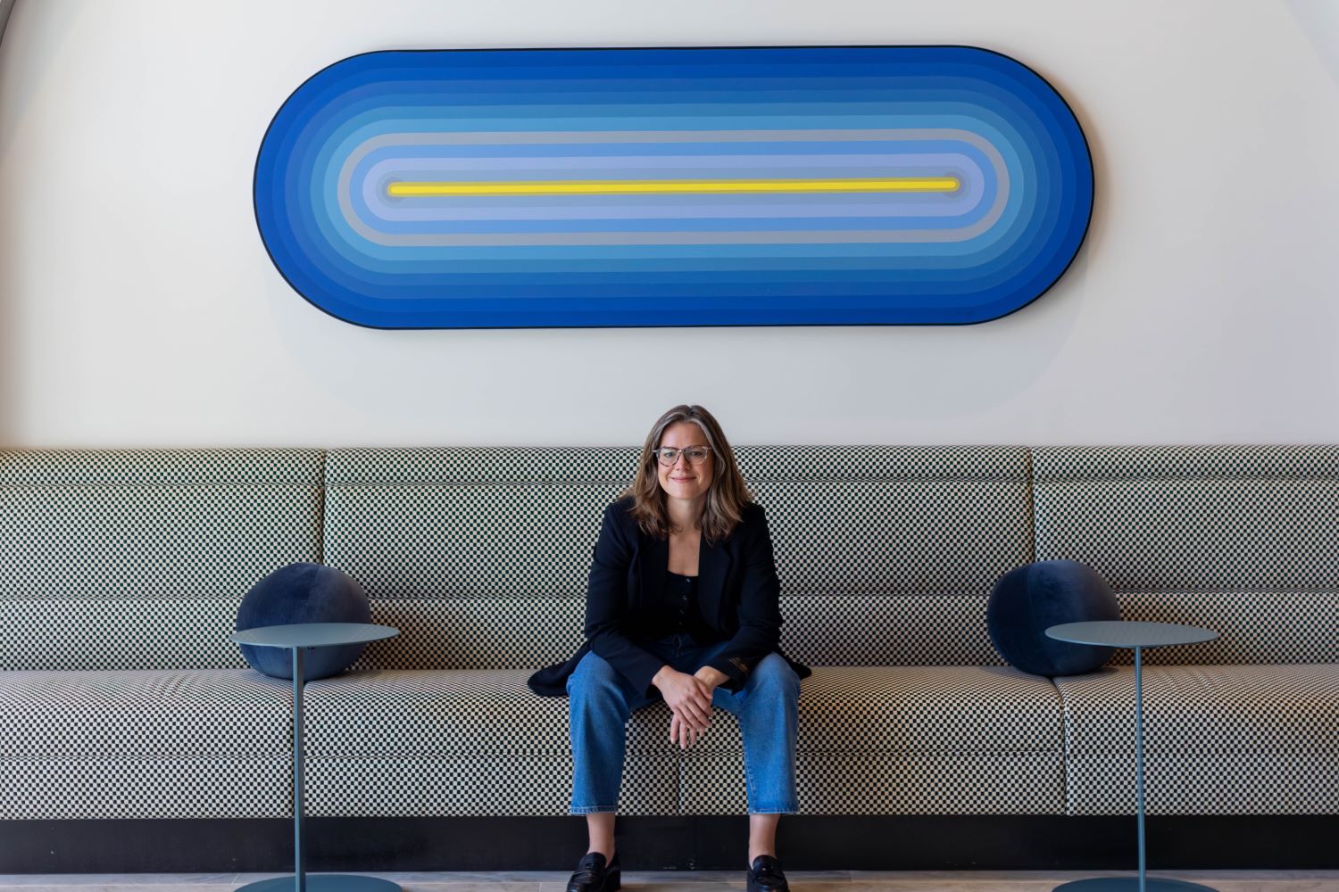 Designer Sheena Brittingham sitting on a bench looking forward with a large blue oval artwork behind her.