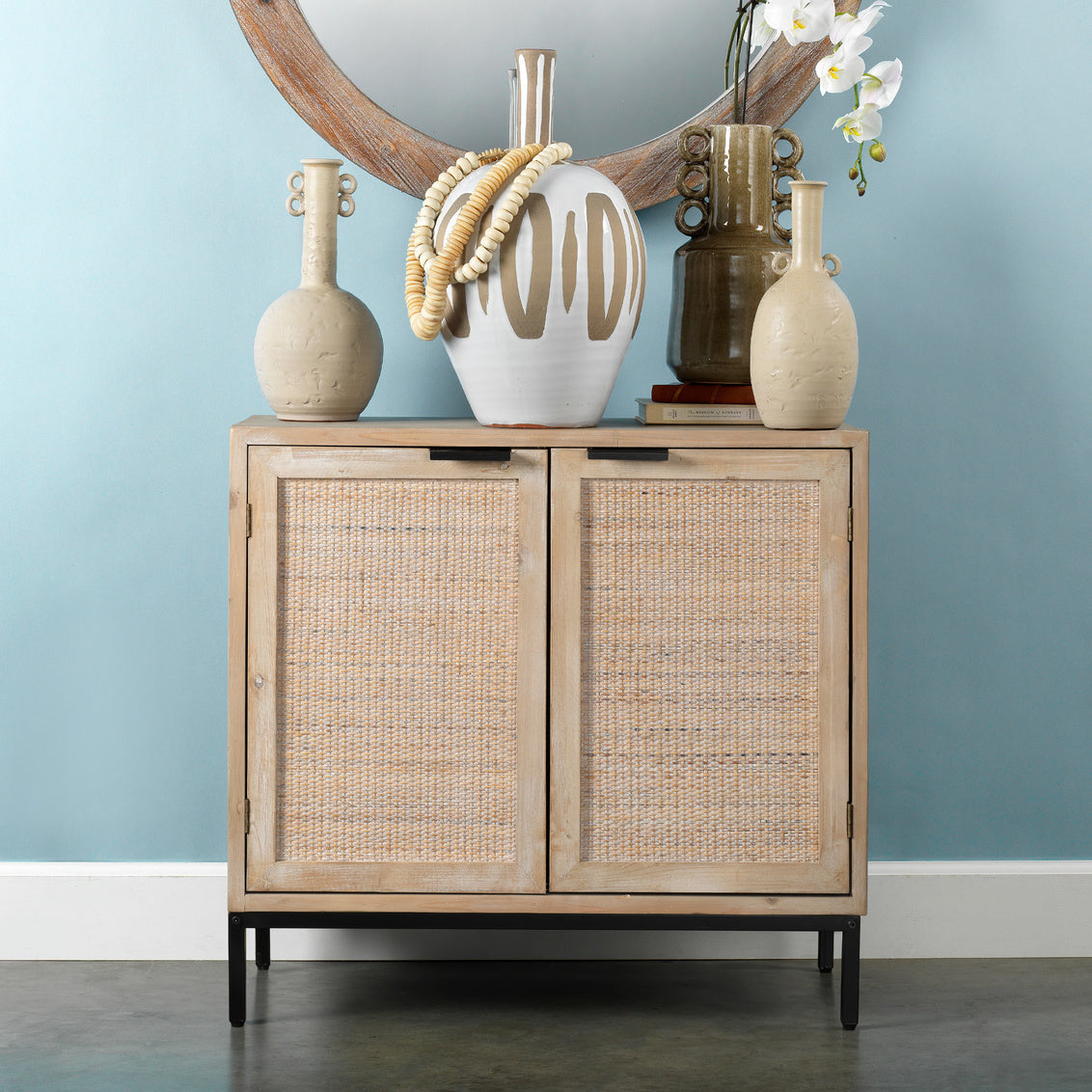 Wooden cabinet with wicker doors against a light blue wall, featuring decorative vases and a mirror.