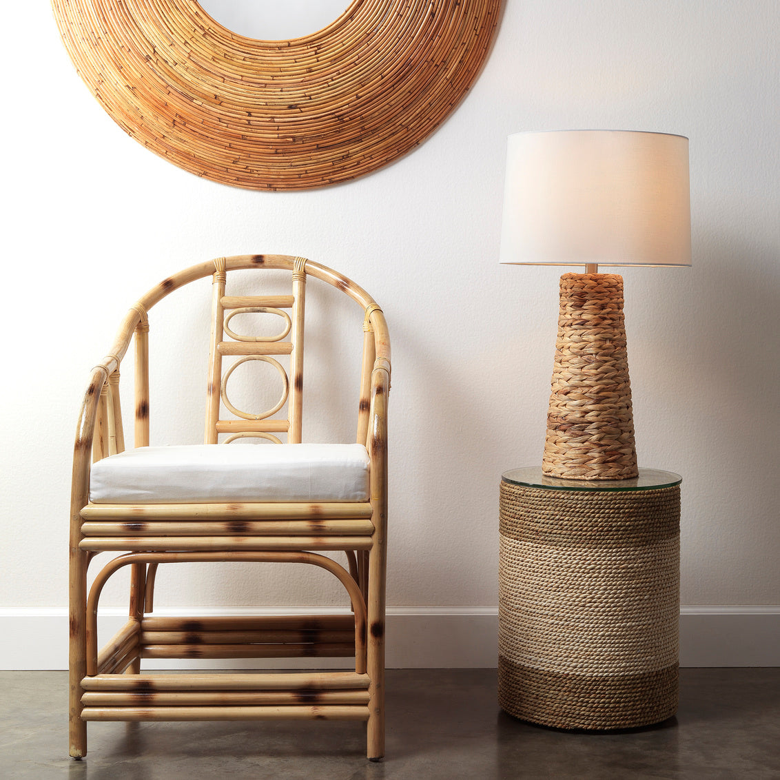 Rattan chair with white cushion next to a rattan side table and lamp against a neutral wall.