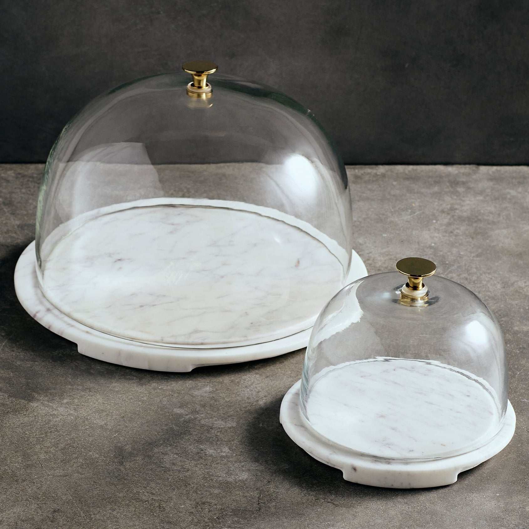 Two glass cake domes with marble bases on a gray surface