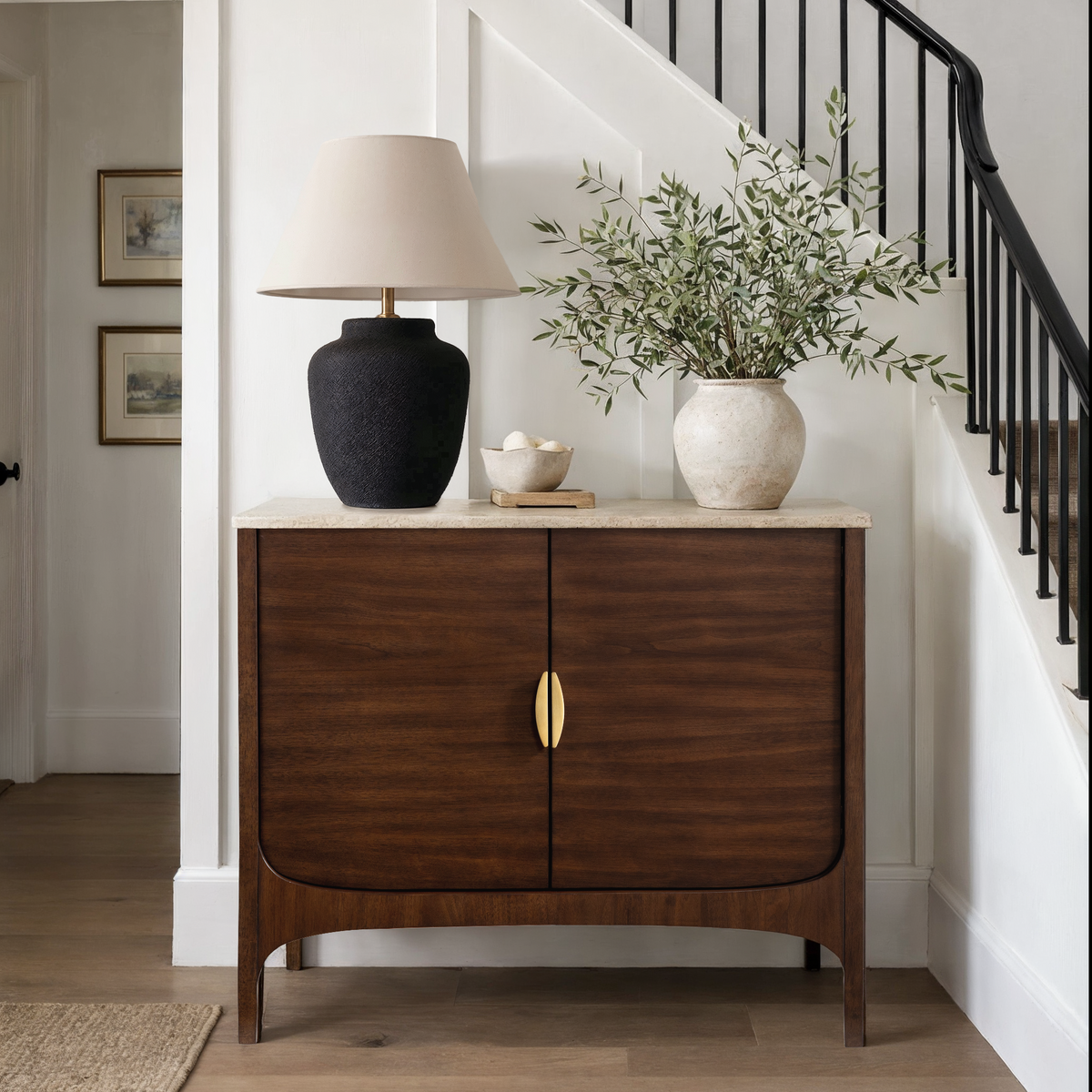 A mid-century modern wooden credenza or sideboard cabinet with two doors and gold-tone handles, styled in a home entryway under a staircase with a lamp and a plant.