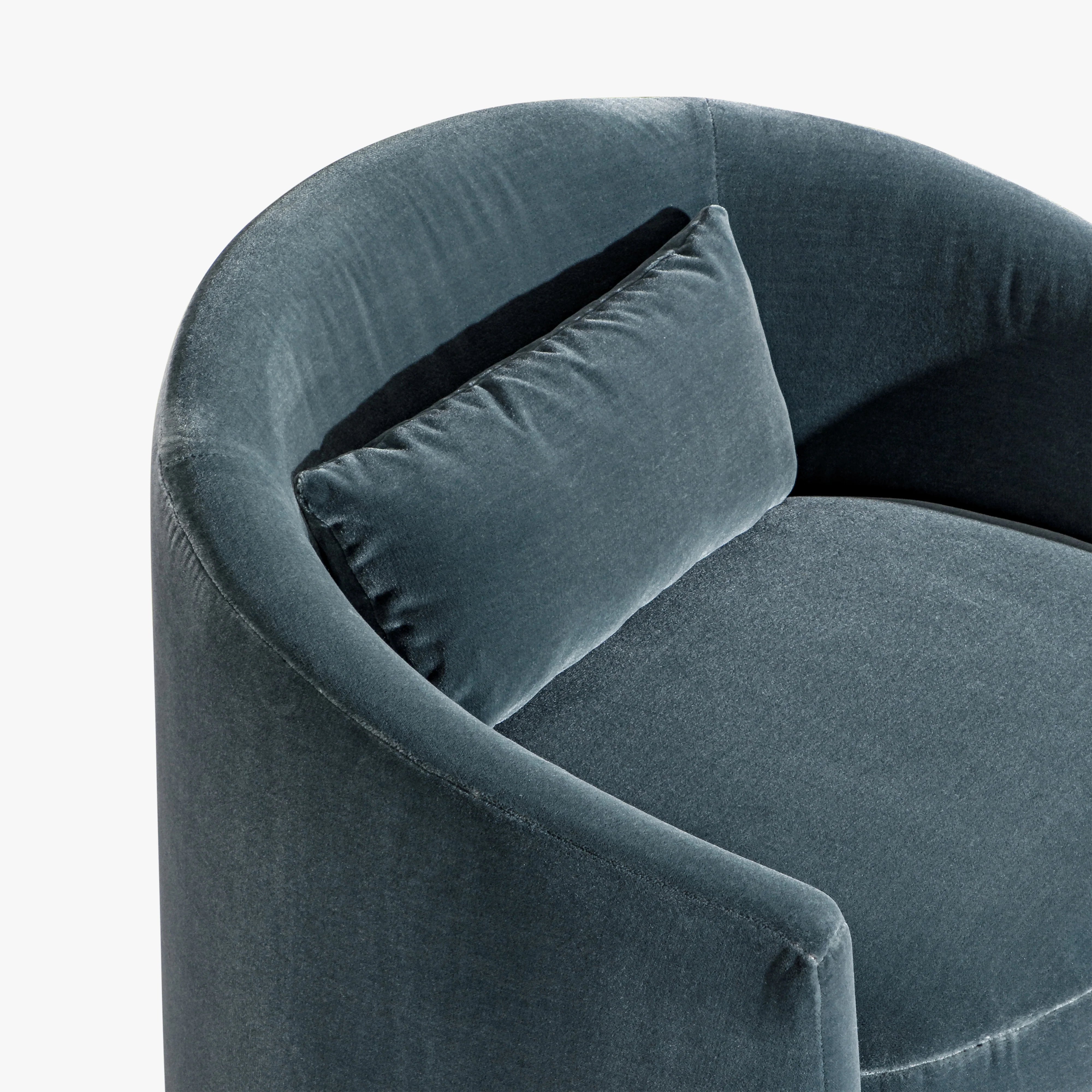 A close-up of a curved upholstered armchair in dark blue velvet with a matching square throw pillow, shown against a plain white background.