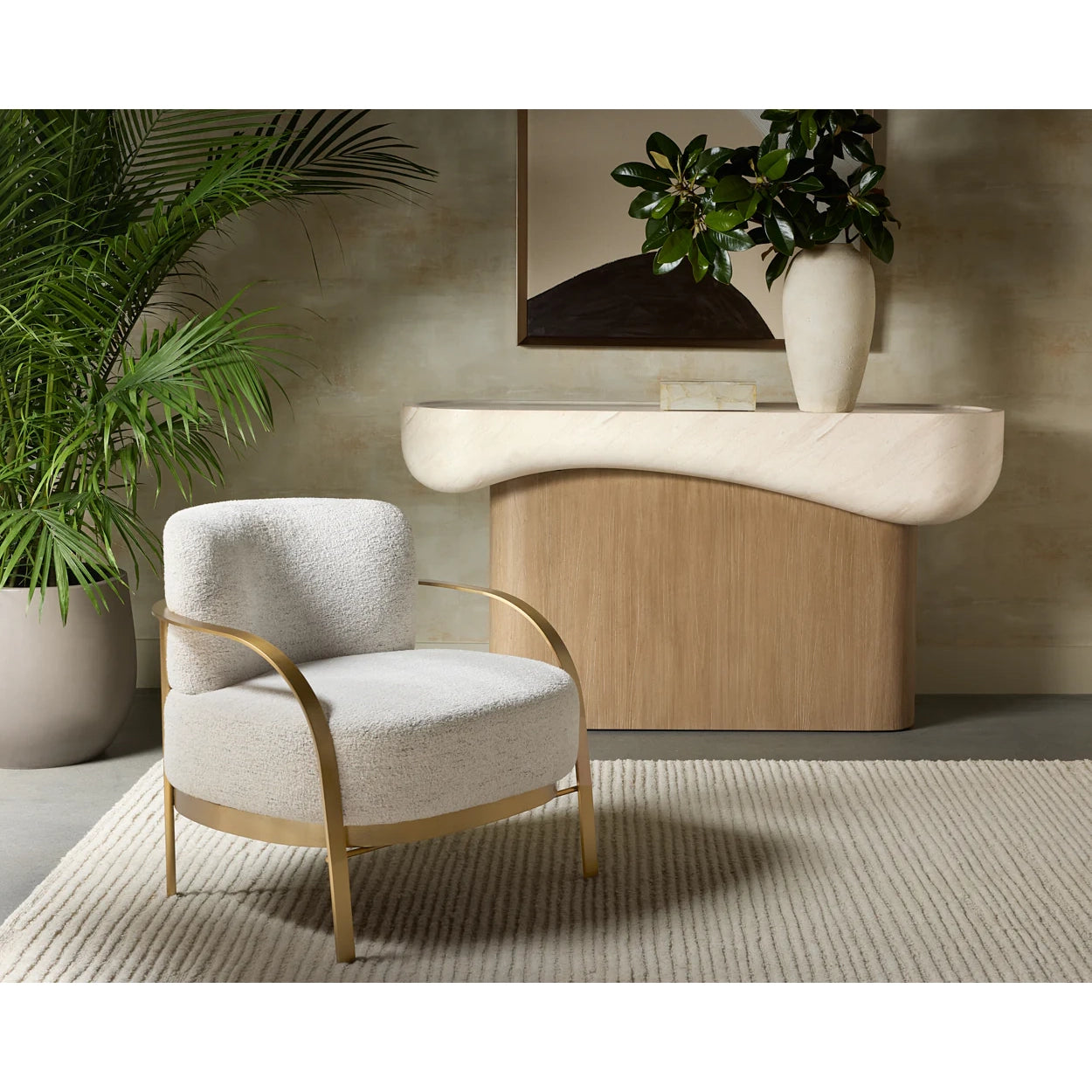 A modern upholstered accent chair with textured off-white fabric and curved gold metal arms and frame, placed in a styled interior setting with a console table, vase, and potted plant.