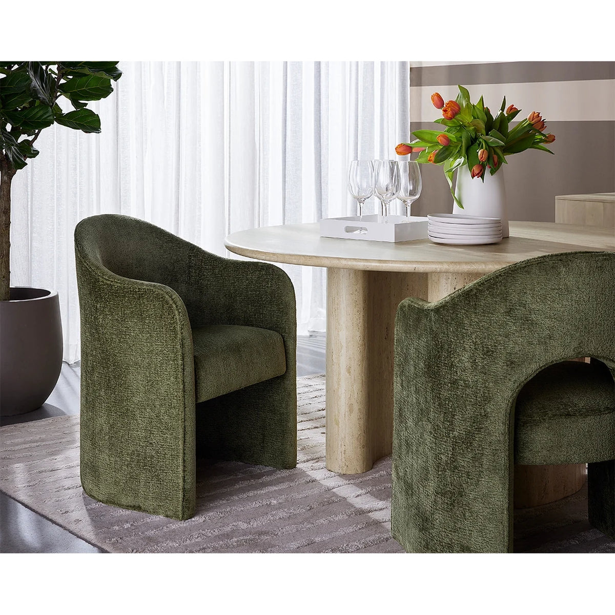Two modern upholstered dining chairs in textured olive green fabric, shown in a styled dining room setting with a light wood table, white plates, wine glasses, and a vase of orange flowers.