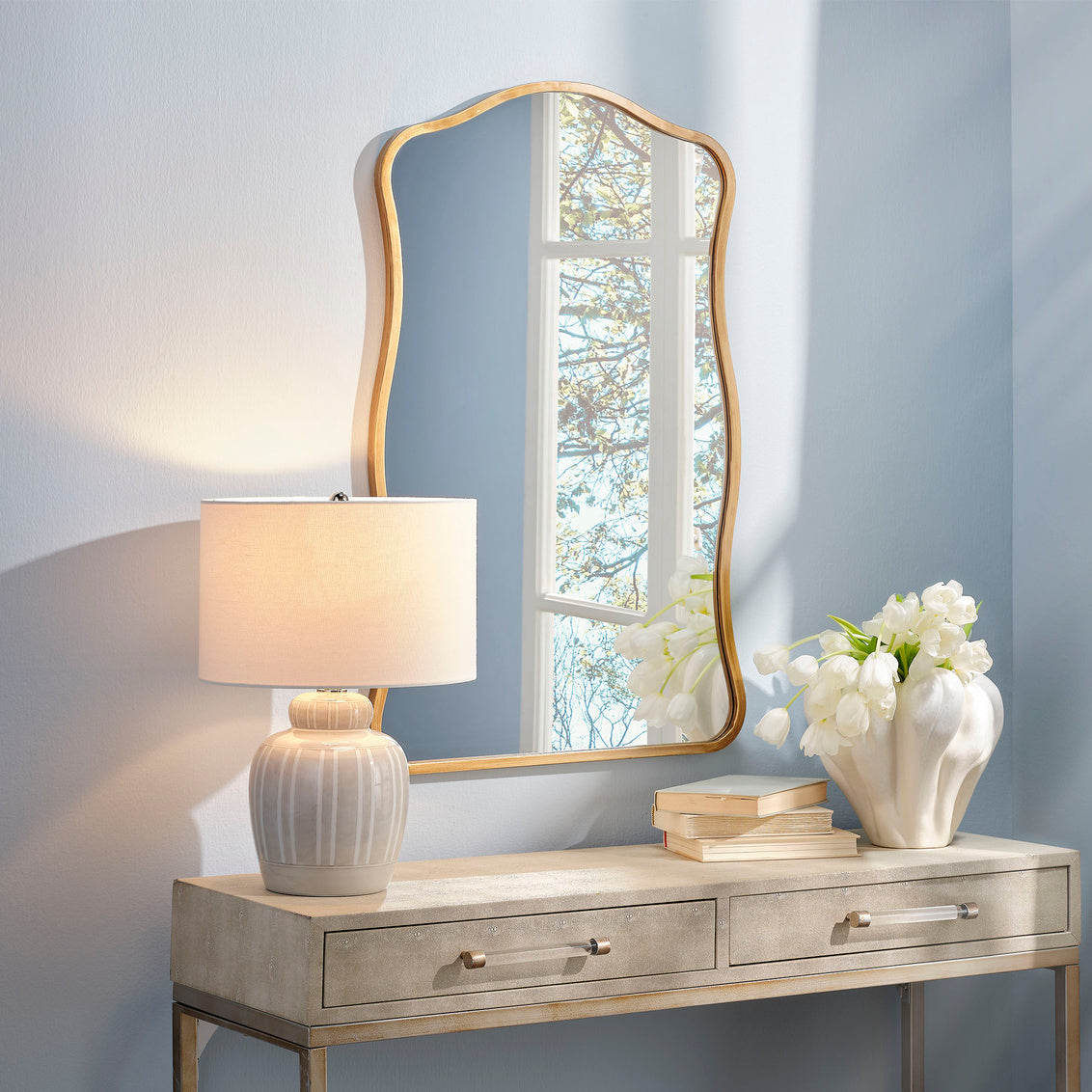 Console table with brass wall mirror, lamp, and flowers against a light blue wall