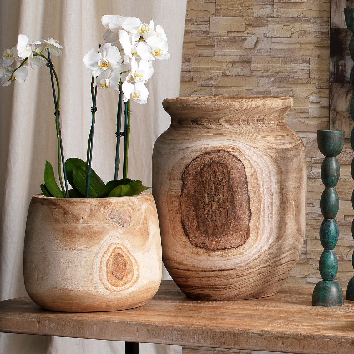 Two wooden vases with a plant on a wooden surface against a stone wall.