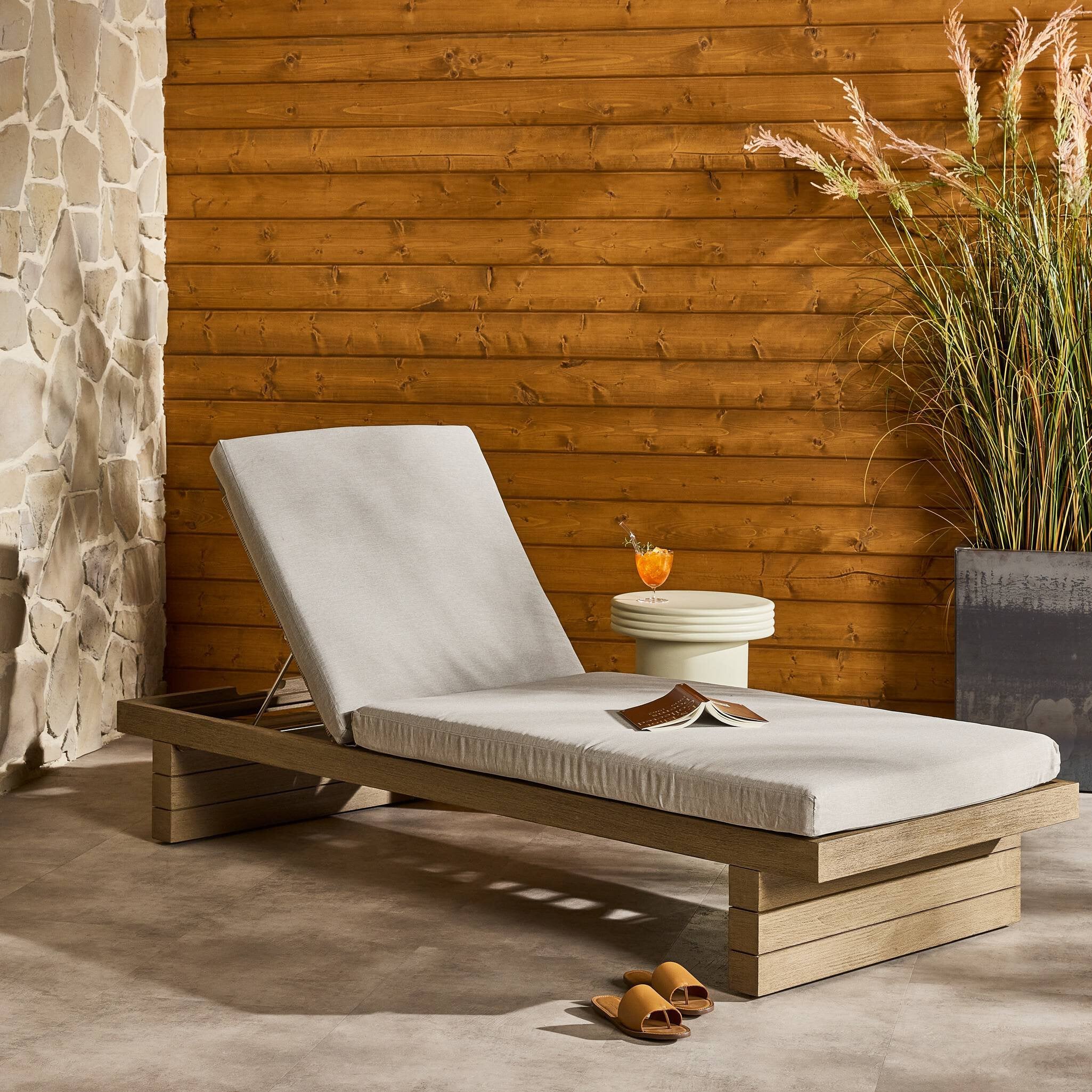 Wooden outdoor lounge chair with white cushion against a wooden wall with a stone fireplace.