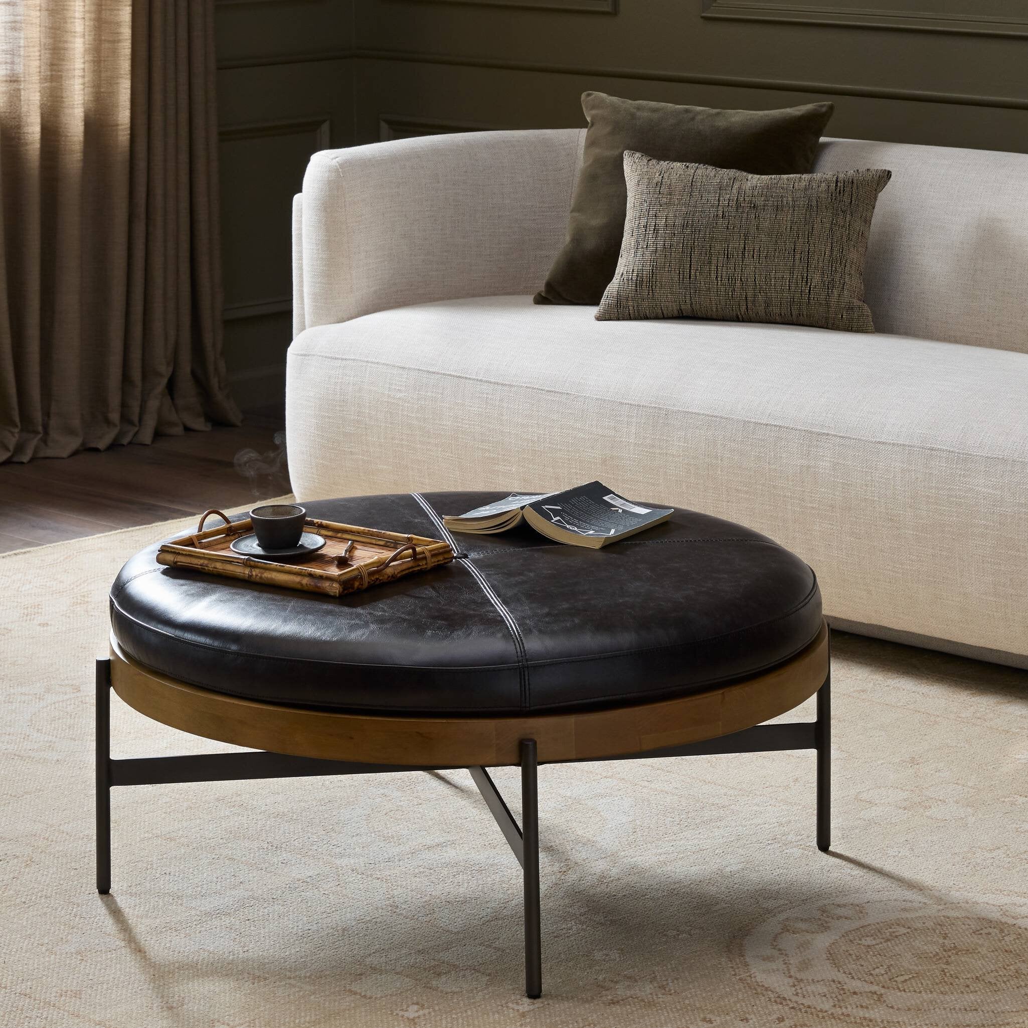 Round black leather ottoman with a book and tray in front of a beige sofa.