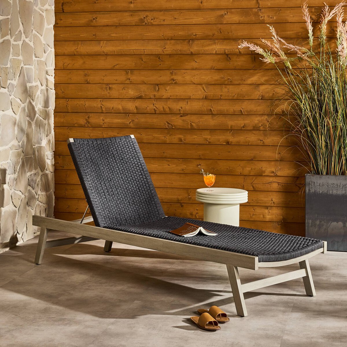 Black woven lounge chair in a patio setting with a wooden wall and stone wall in the background.