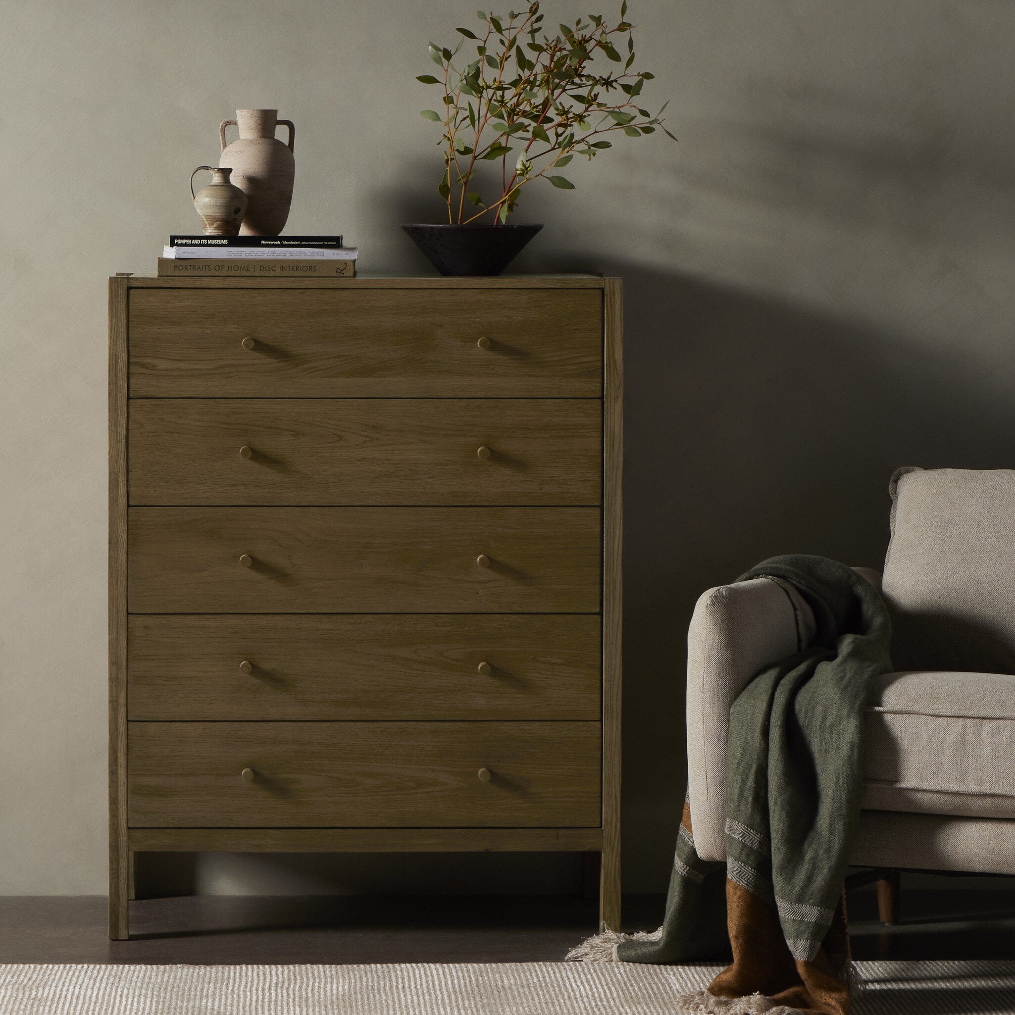 Wooden dresser with decorative items next to a gray armchair in a room with a neutral color scheme.