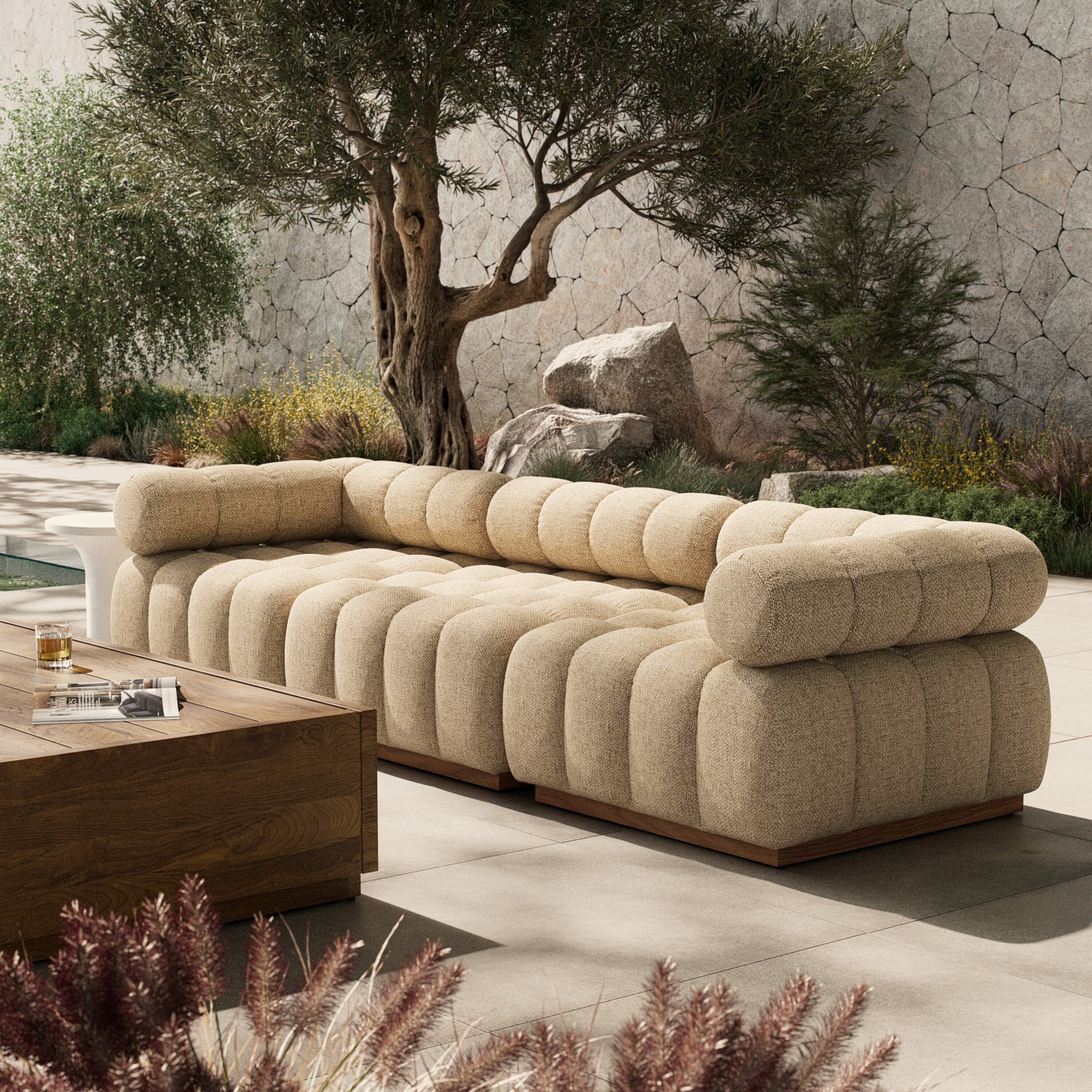 Beige sectional sofa in an outdoor setting with a stone wall and tree in the background.