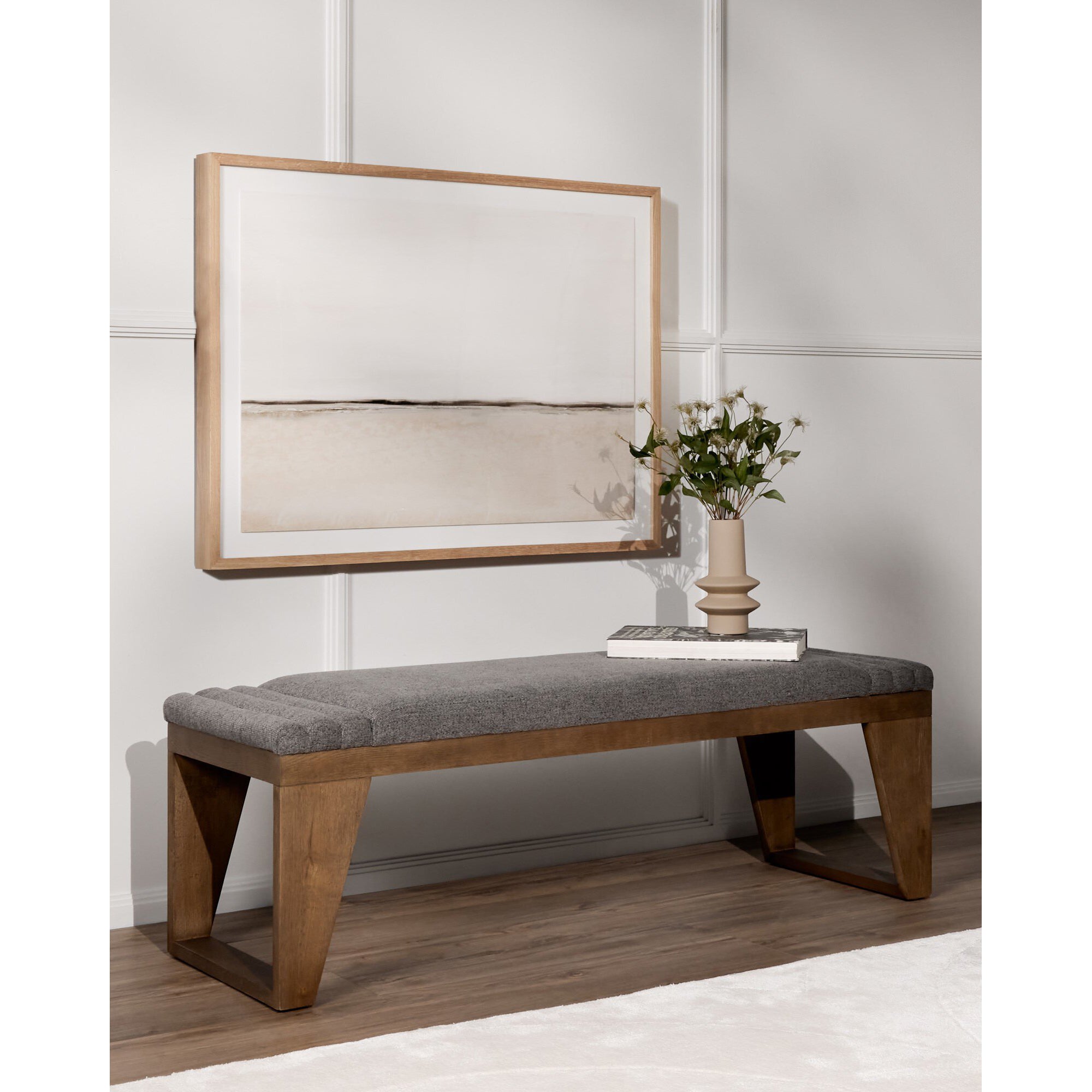 Large framed artwork of a minimalistic landscape hanging above a wooden bench with gray upholstery in a modern room