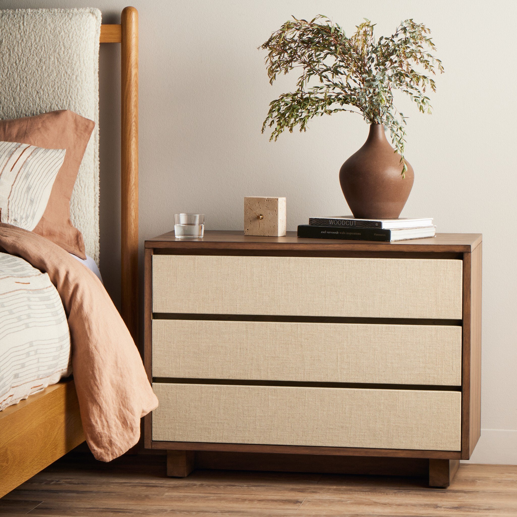 Wooden nightstand with beige fabric drawers next to a bed with brown bedding.