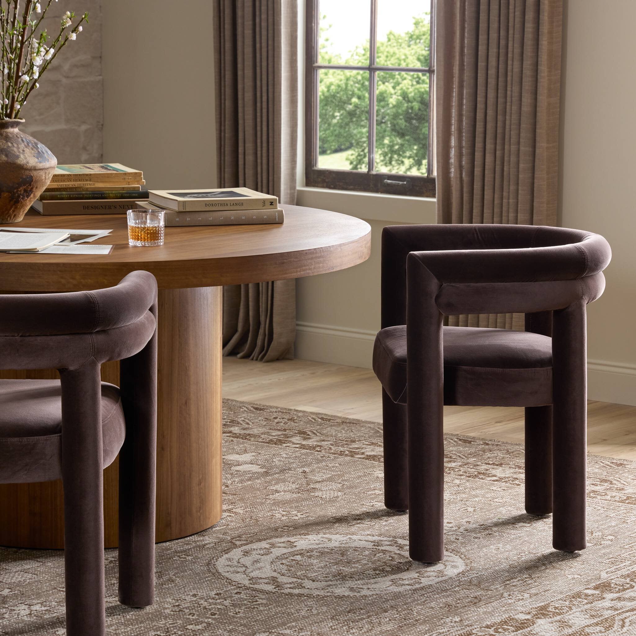 Round wooden dining table with two dark purple chairs in a room with large windows and curtains.