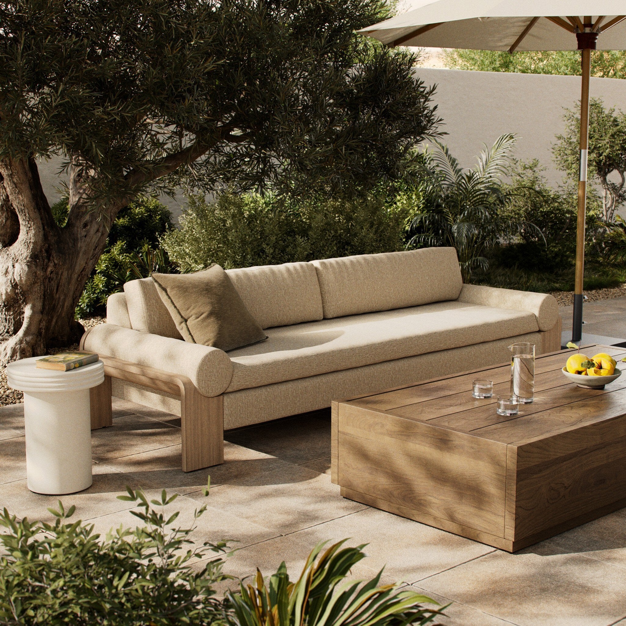 Outdoor patio setting with beige sofa, wooden coffee table, and umbrella in a garden.