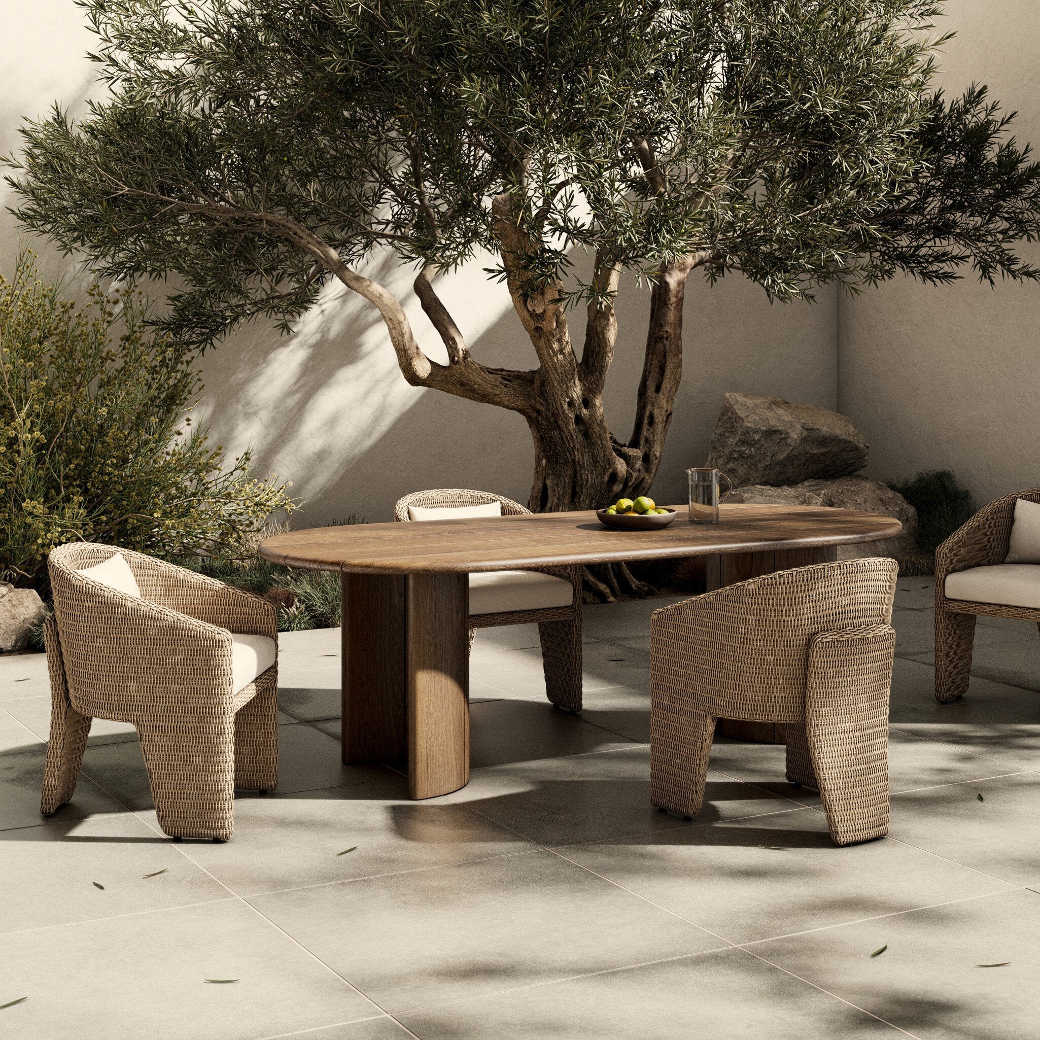 Outdoor dining area with wooden table and wicker chairs under a large tree.
