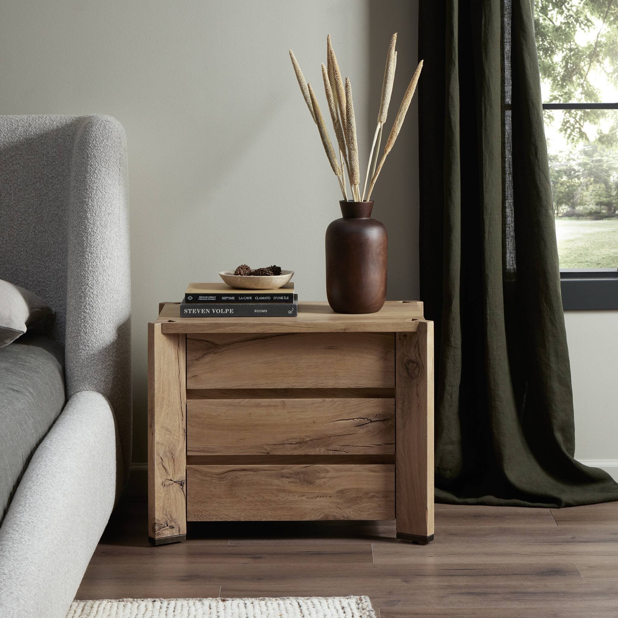 Wooden nightstand with a vase and books next to a gray sofa in a room with a window and curtain.