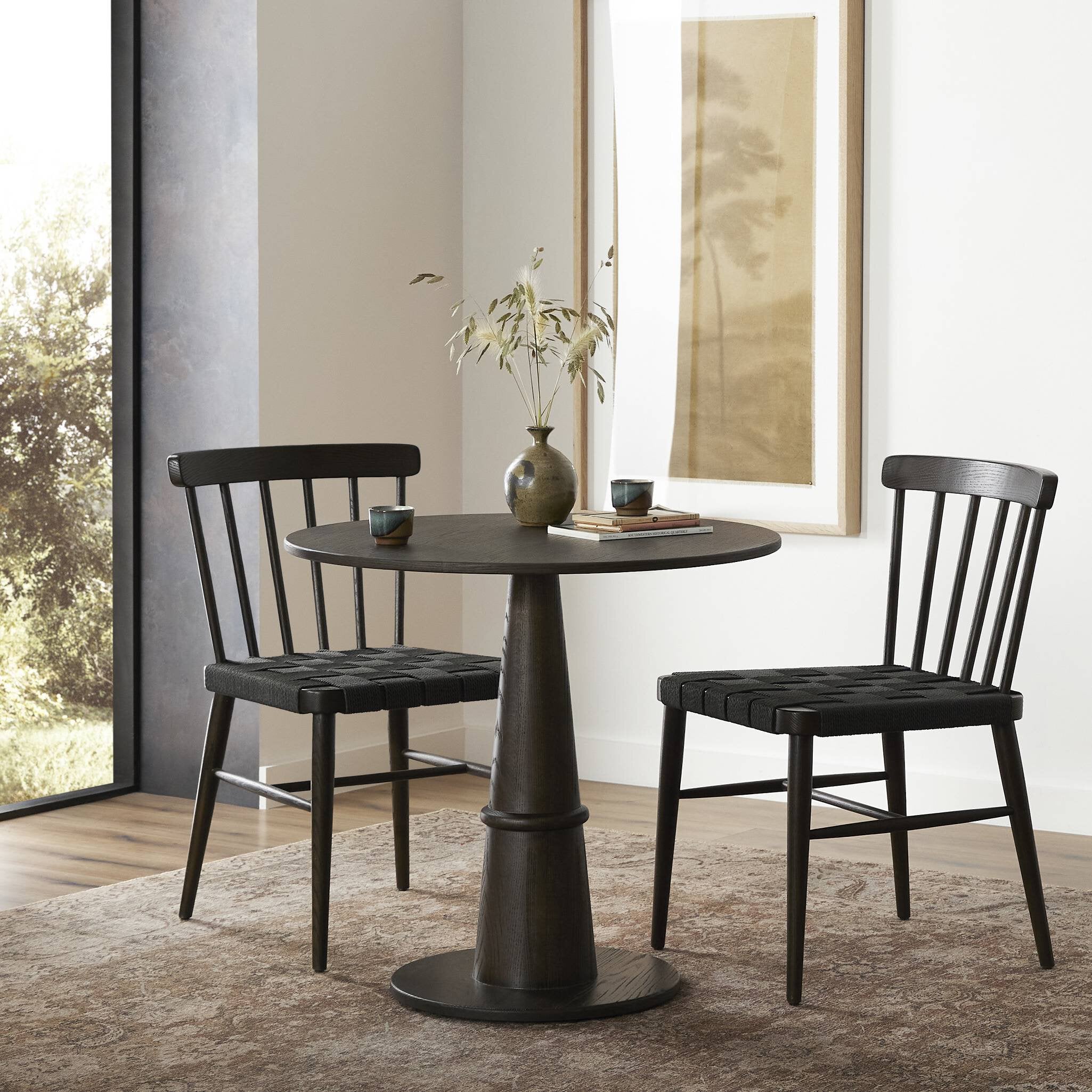 Dining set with a round black table and two matching chairs in a room with large windows.