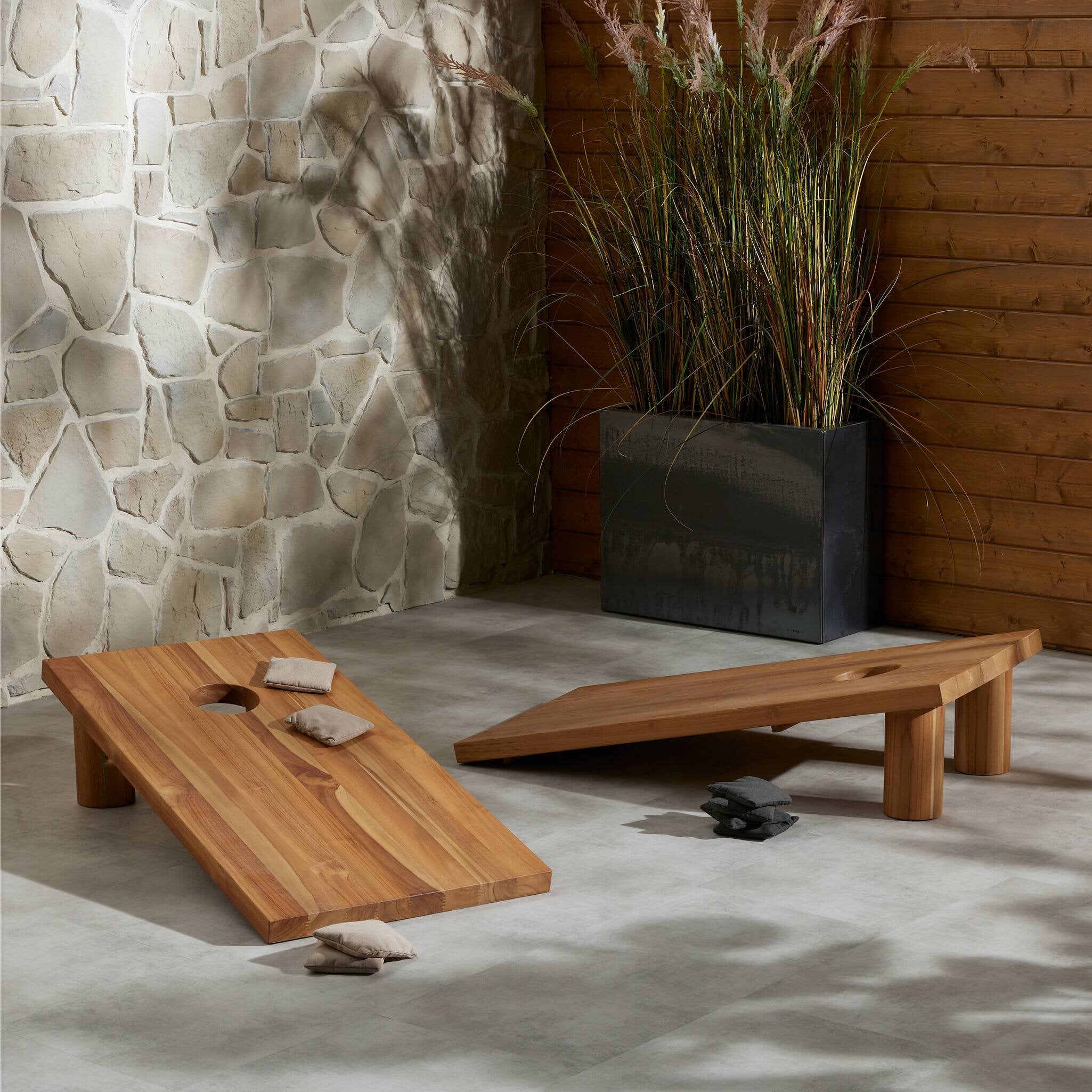Wooden cornhole tables against a stone wall with decorative plants.