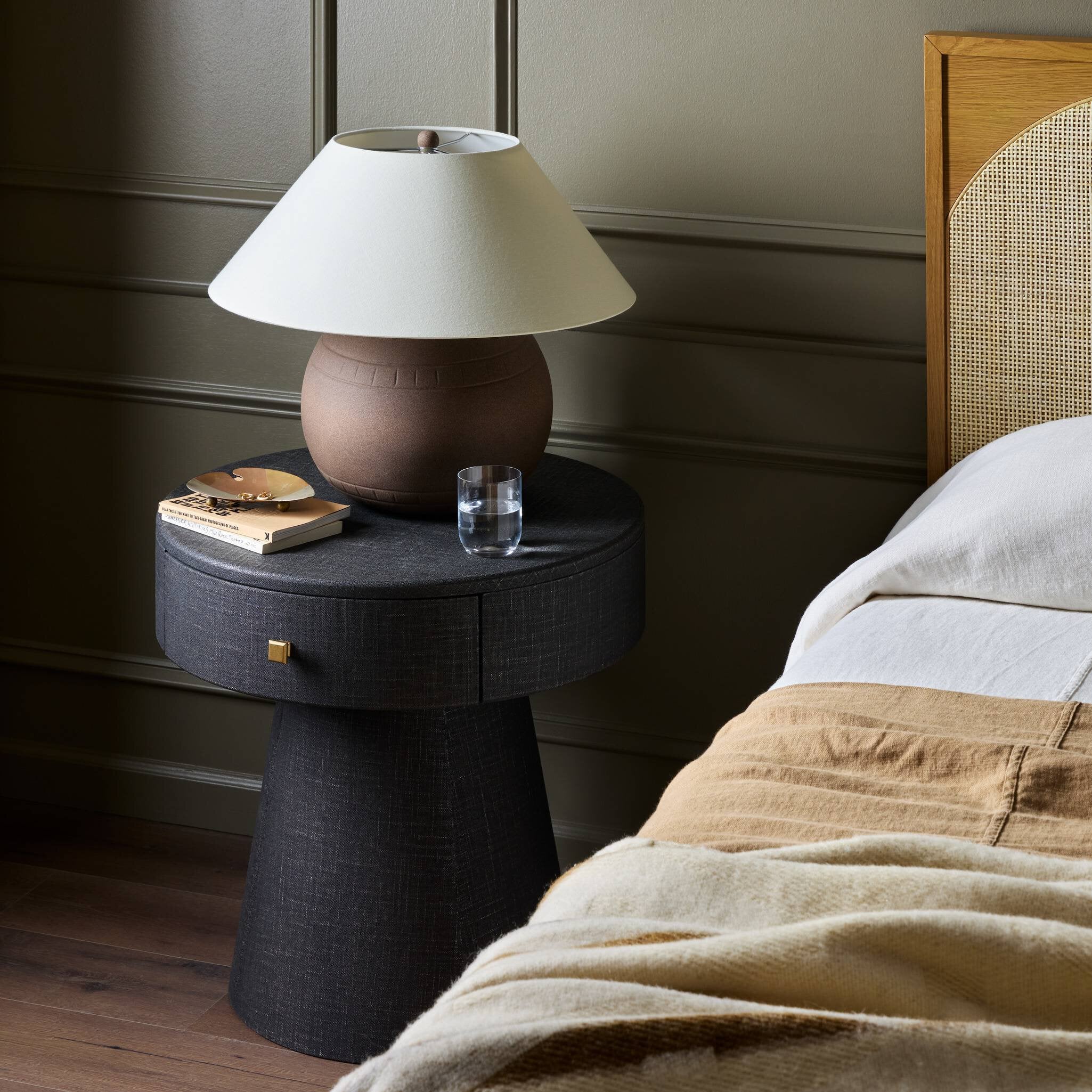 Black side table with a lamp and glass next to a bed with beige bedding.