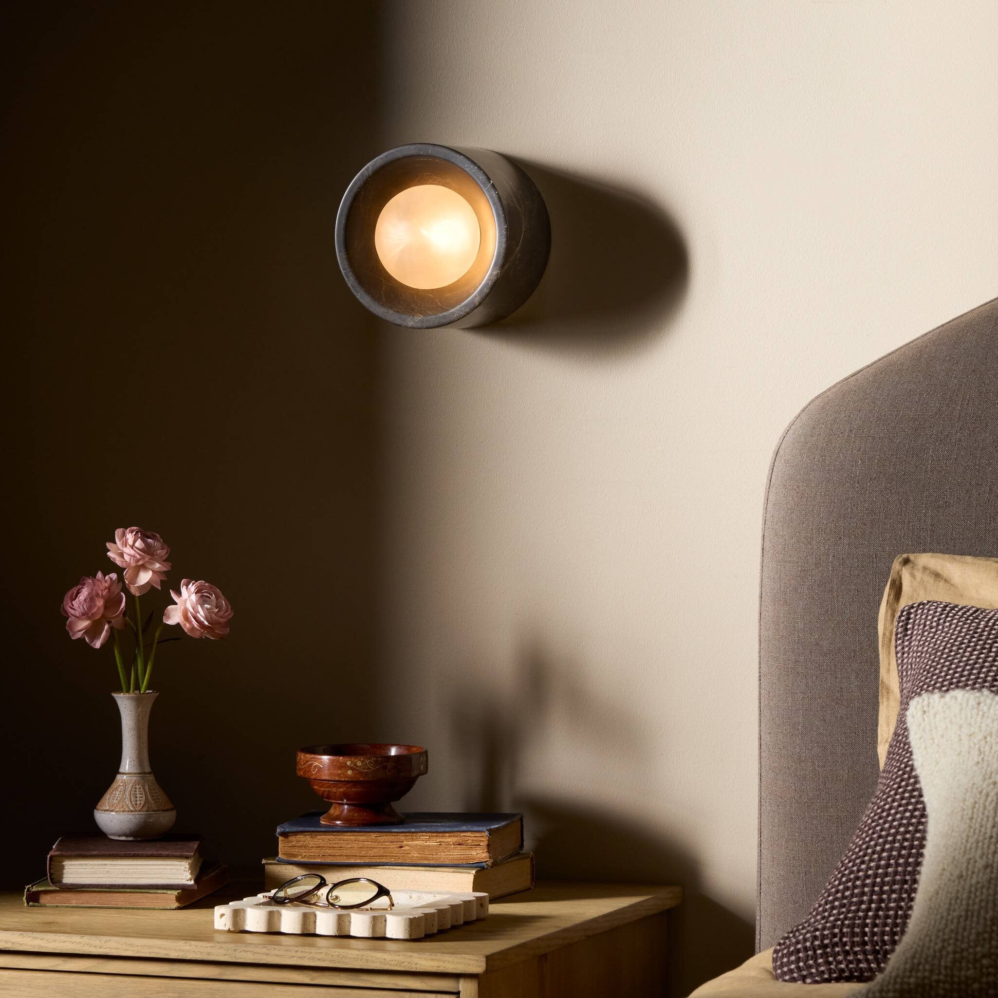 Wall-mounted light fixture casting a warm glow on a wall next to a wooden nightstand with books and a vase.