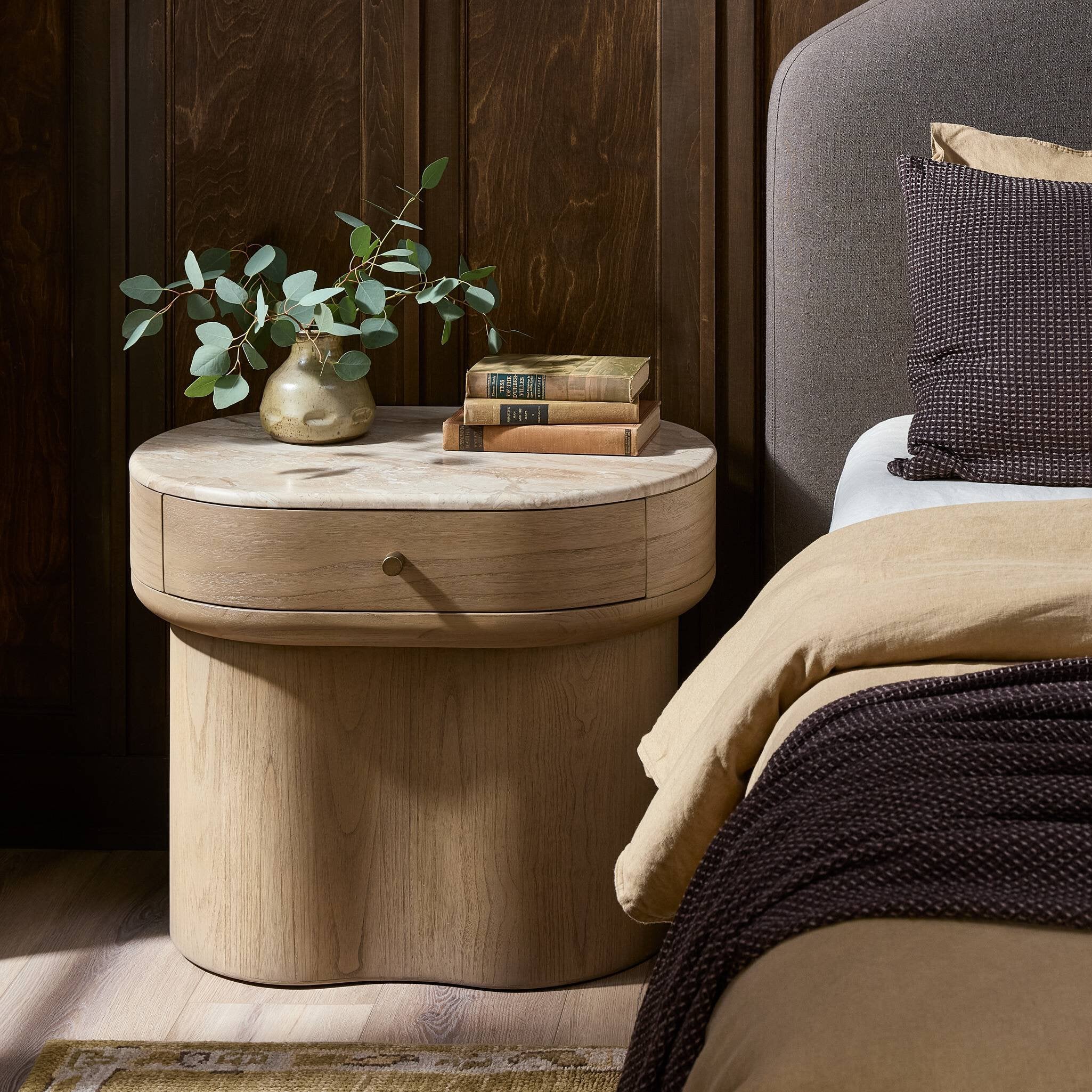 Wooden side table with a plant and books next to a bed with pillows and blankets.