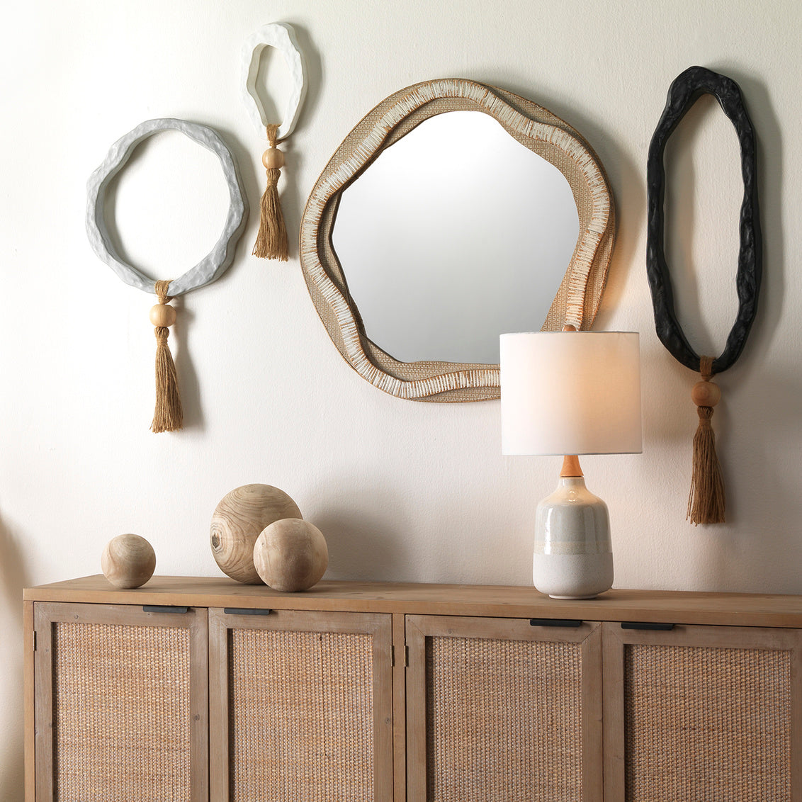 Decorative mirrors with tassels and a lamp on a wooden console against a white wall.