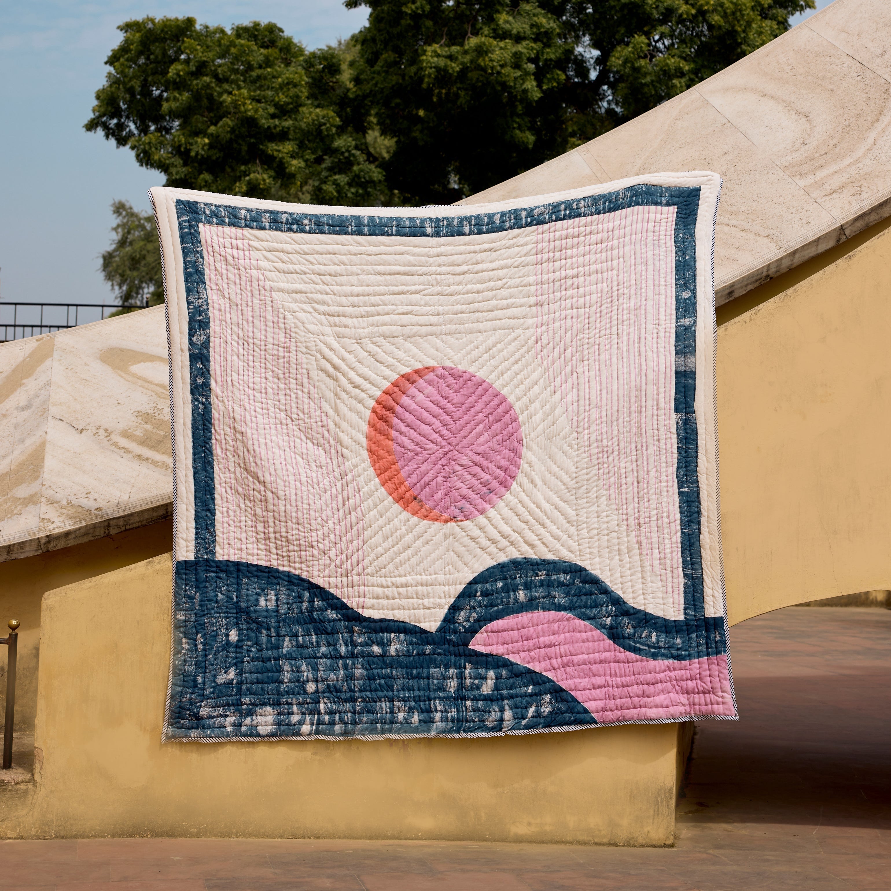 Quilt with a pink sun design draped over a building exterior.