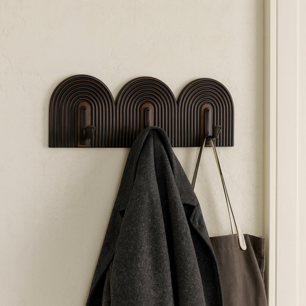 Decorative wall hook with three hooks holding a coat and a bag on a beige wall.