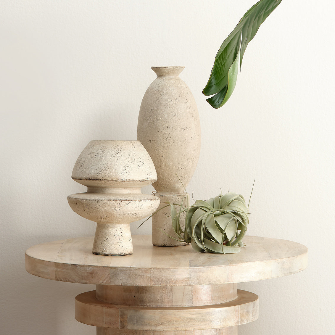 Two ceramic vases on a round wooden table with a plant on a light background