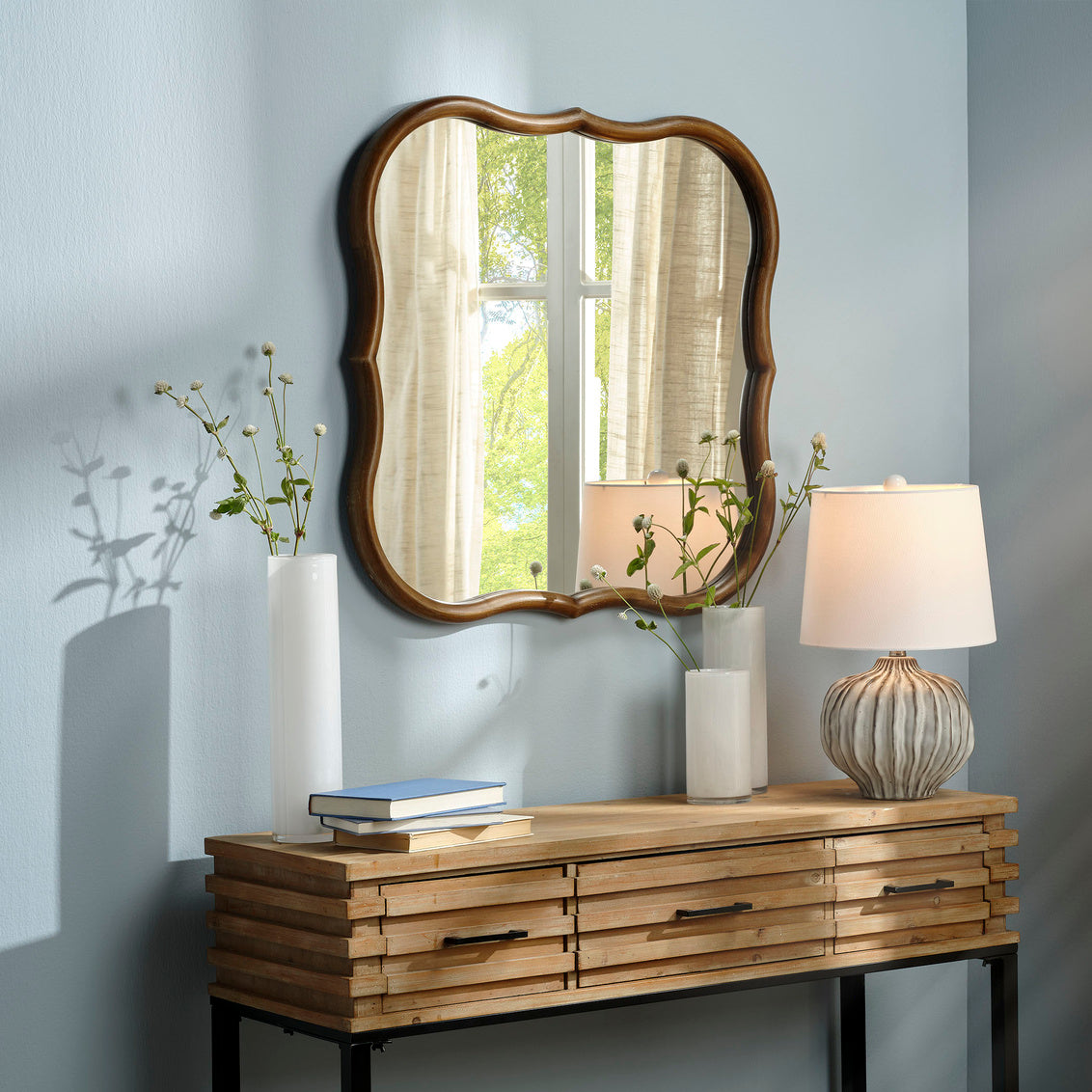 Wooden console table with mirror, lamp, and decorative items against a light gray wall.