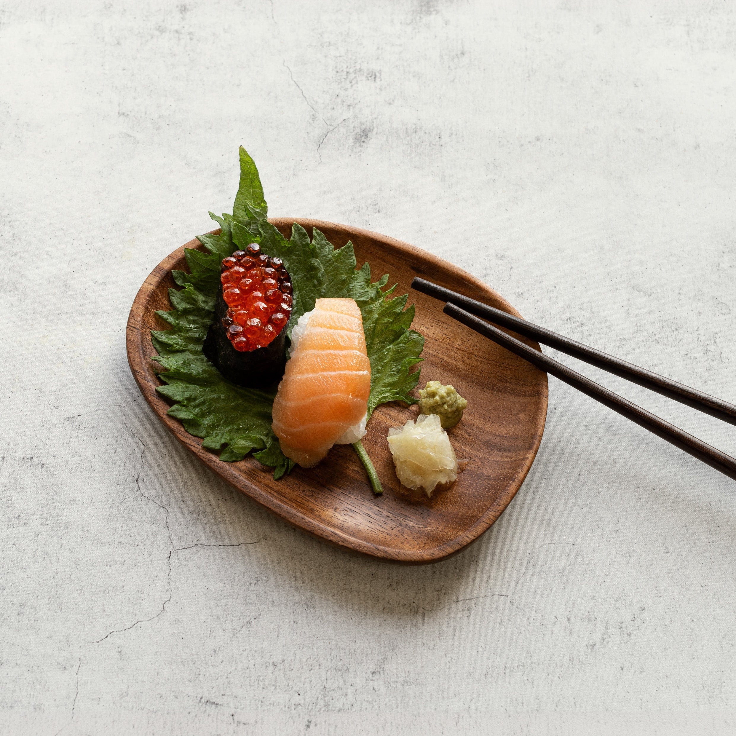 Sushi on a wooden plate with leaves and wasabi, accompanied by chopsticks on a light gray background.