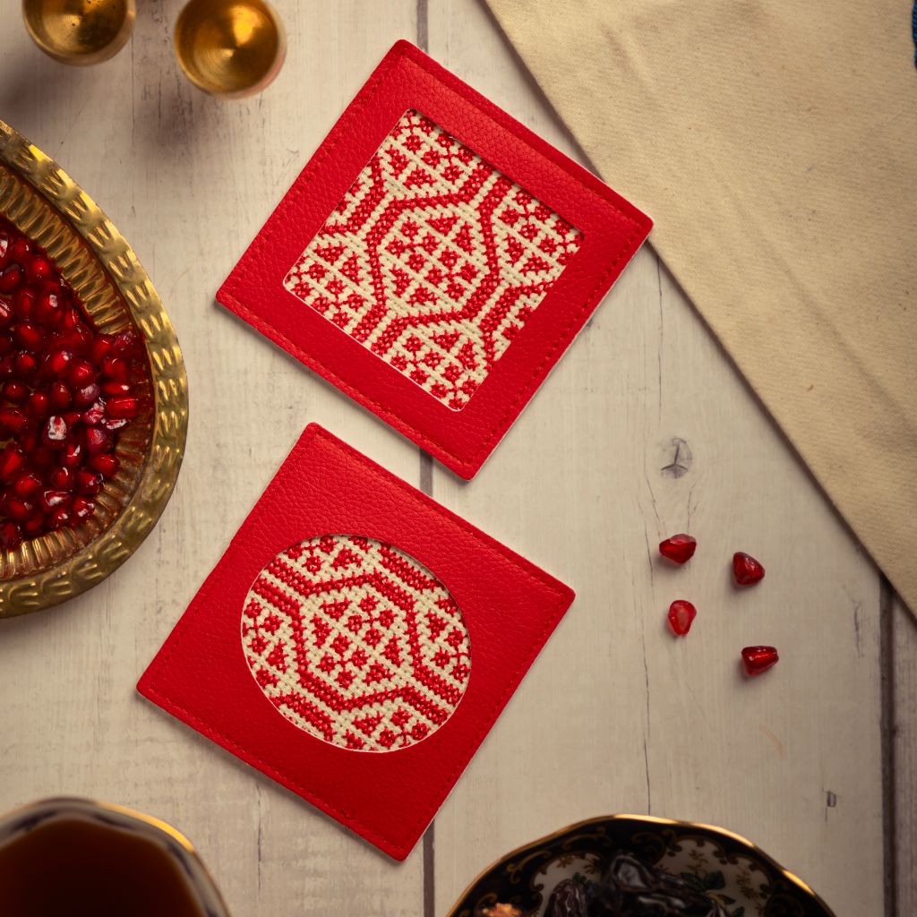 Two red coasters with white patterns on a wooden surface with pomegranate seeds.