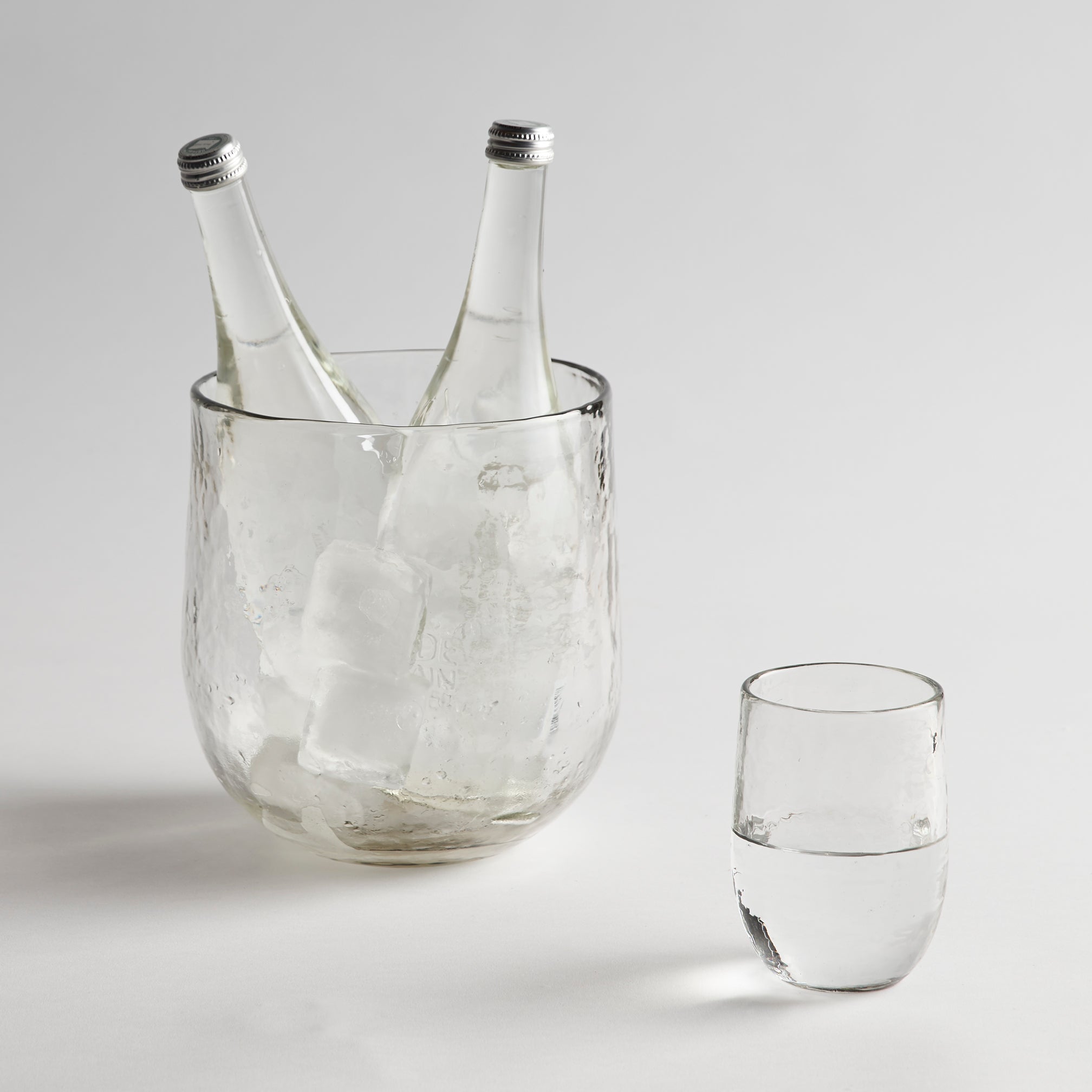 Clear glass ice bucket with two bottles and a glass of water on a light gray background