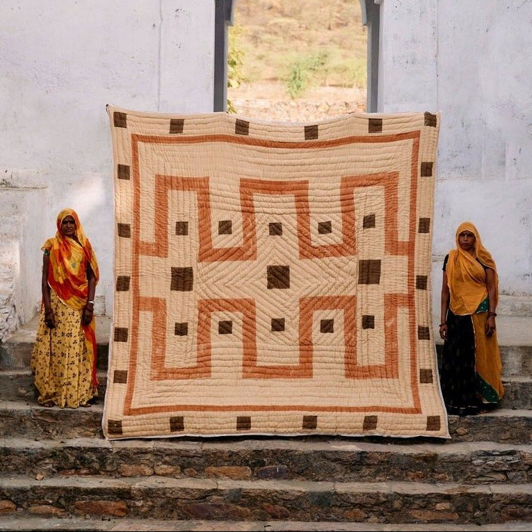 Two women standing on steps with a large decorative quilt between them, against a white wall with an archway.