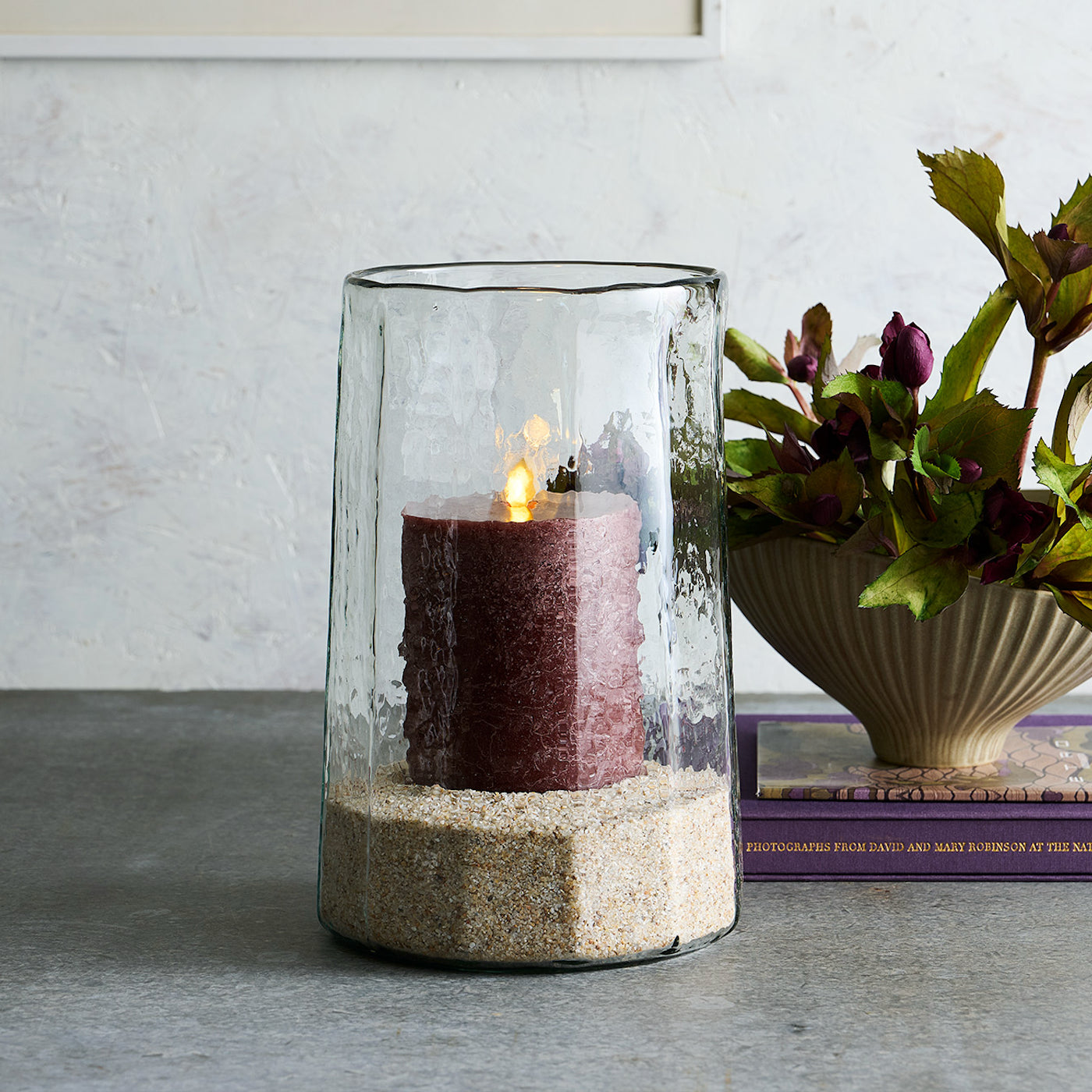 Candle in a glass holder with sand on a table, next to a plant and books.