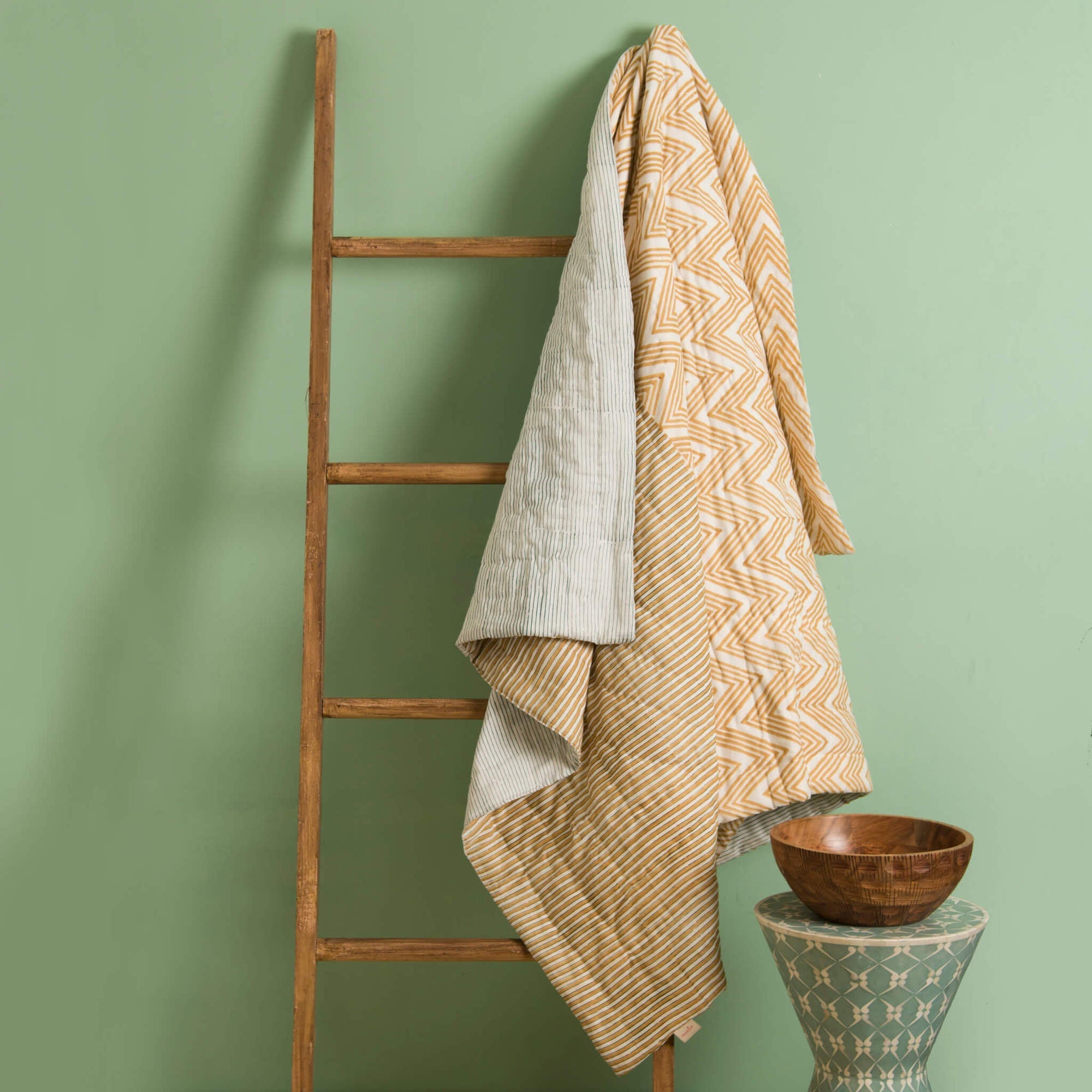 Wooden ladder with yellow patterned throw blanket against a green wall, wooden bowl on a small table.