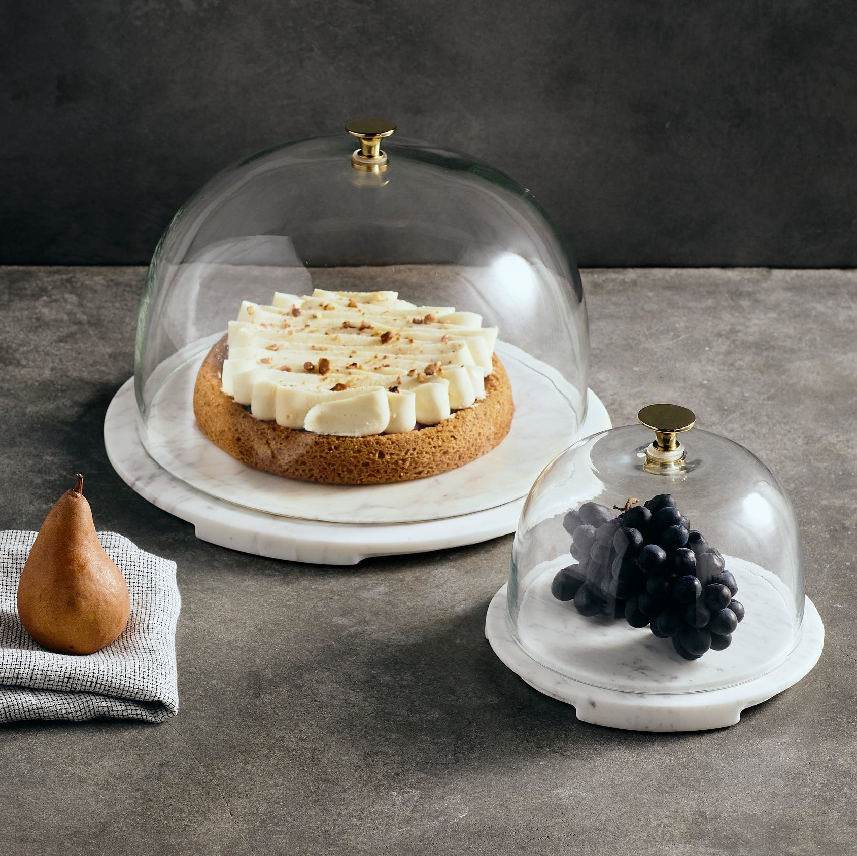 Two glass domes on marble bases with a cake and grapes respectively, on a dark surface.
