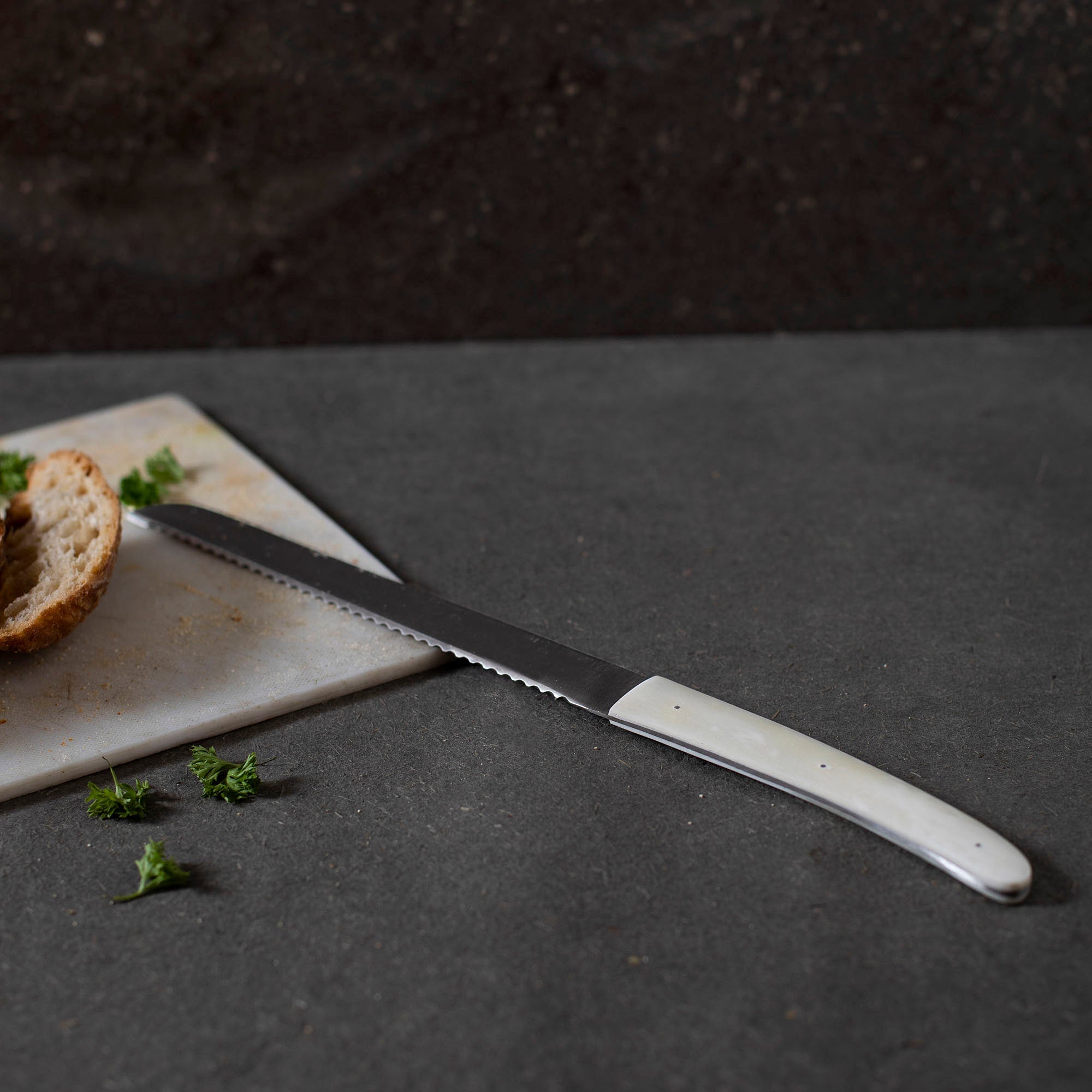 Bread knife with a white handle on a dark surface next to a slice of bread and herbs.