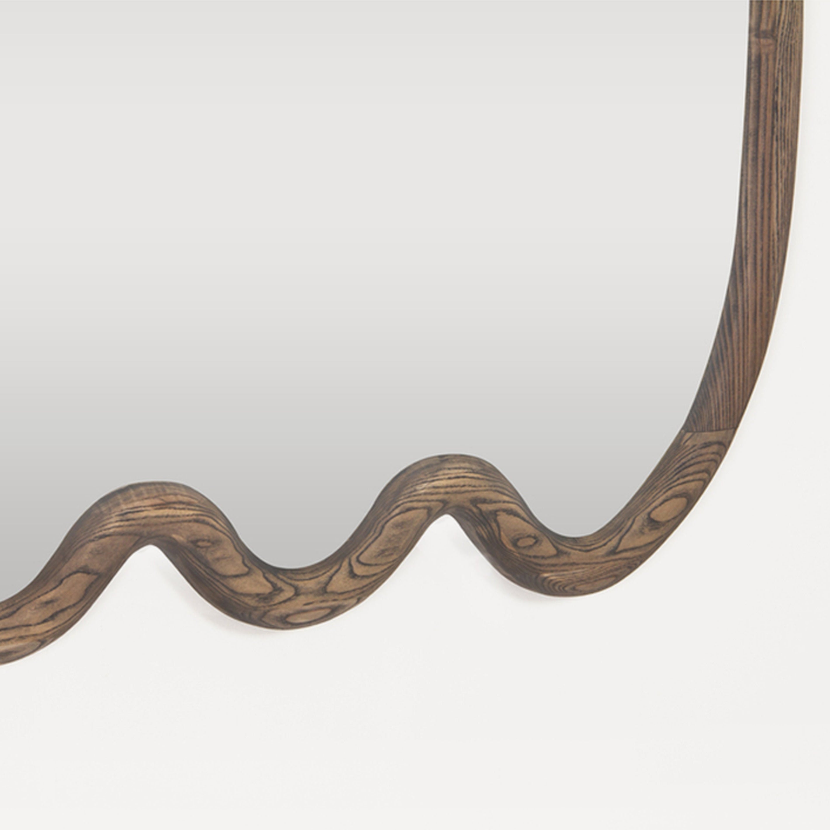 A close-up of a decorative wall mirror with a wavy, undulating wooden frame in a dark brown finish, set against a plain light gray background.