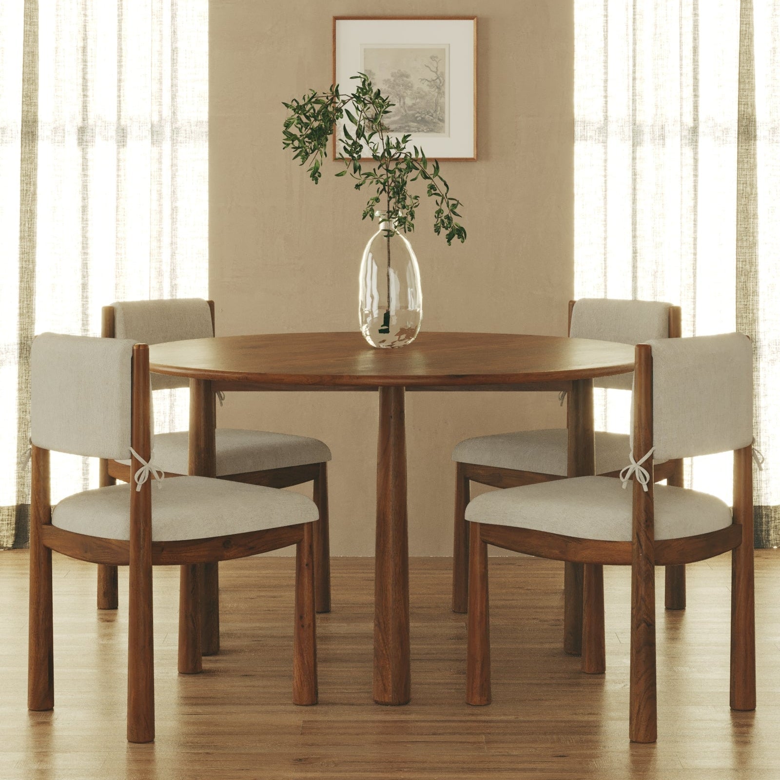 A round wooden dining table with four upholstered chairs in a warm neutral room, styled with a clear glass vase containing green branches on top.