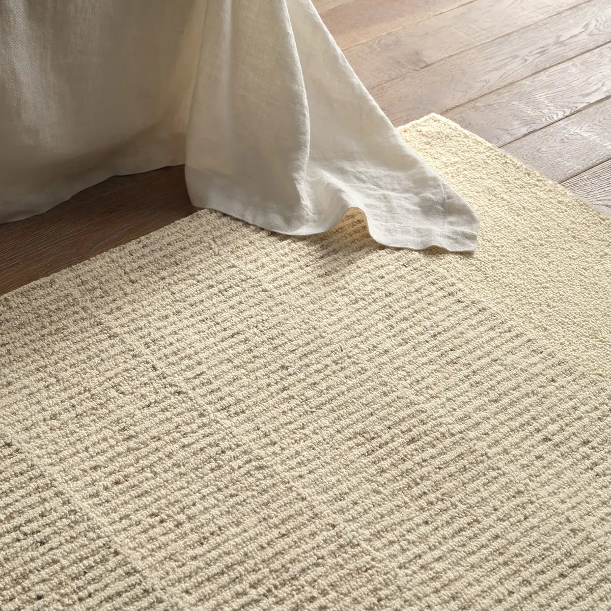 Beige textured rug on a wooden floor with a white fabric corner.