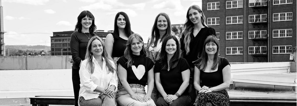A black and white group photograph of eight women posing together outdoors on a rooftop or balcony with buildings in the background.