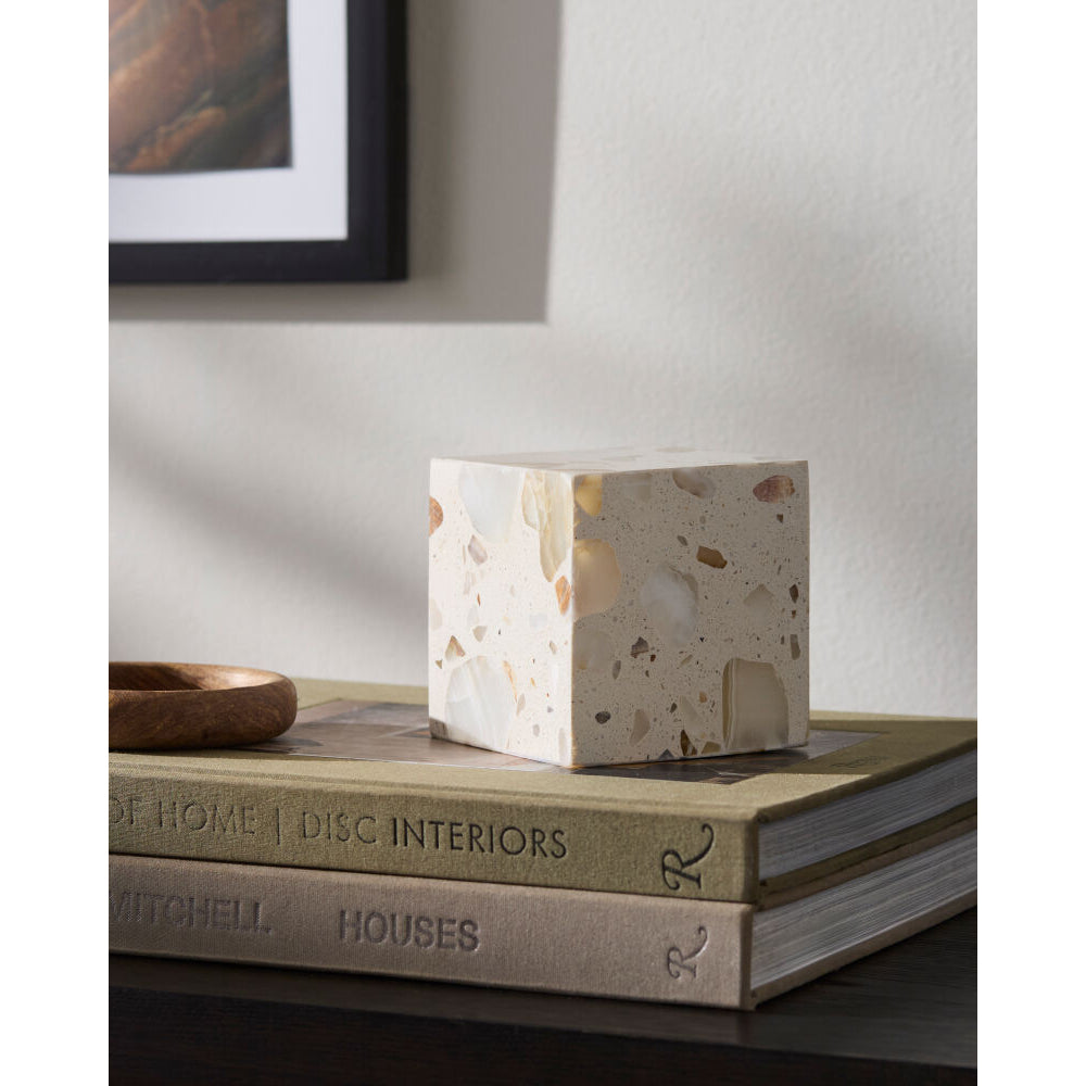 A square terrazzo box in warm beige and cream tones sits on top of two stacked books on a dark wooden surface, with a neutral wall and framed art in the background.