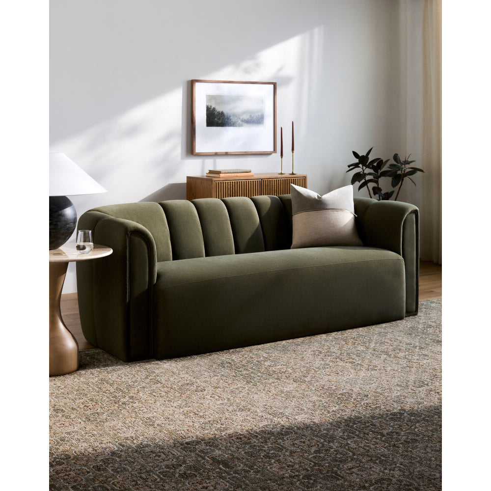 An olive green upholstered sofa with channel tufting on the backrest, styled in a modern living room setting with a neutral throw pillow, side table, lamp, and rug.