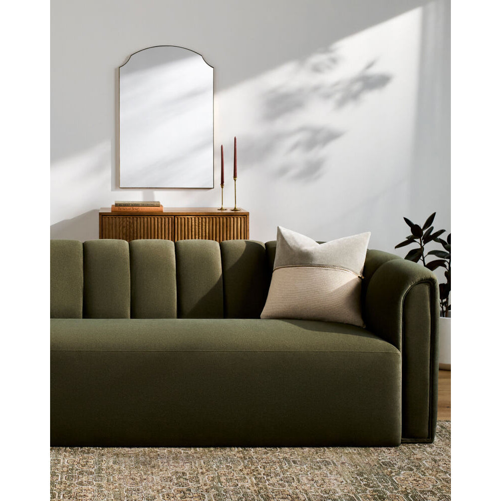 An olive green upholstered sofa with channel tufting and rolled arms, styled in a modern living room setting with neutral throw pillows, a wooden console, a wall mirror, and soft window light casting shadows on the wall.