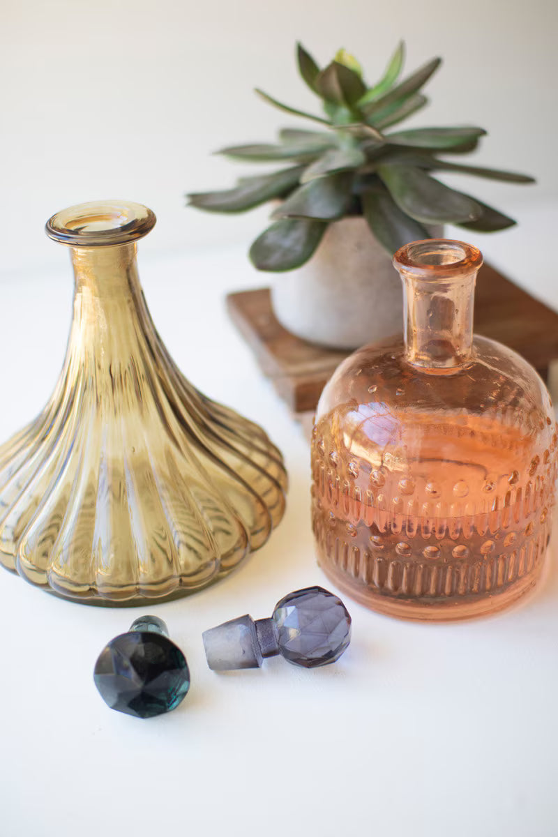 Two glass bottles with decorative caps on a white surface with a plant in the background