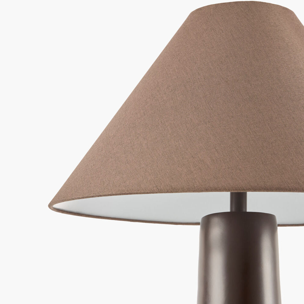 A close-up of a table lamp with a tapered fabric shade in a warm taupe-brown color and a dark metallic cylindrical base, shown against a plain white background.