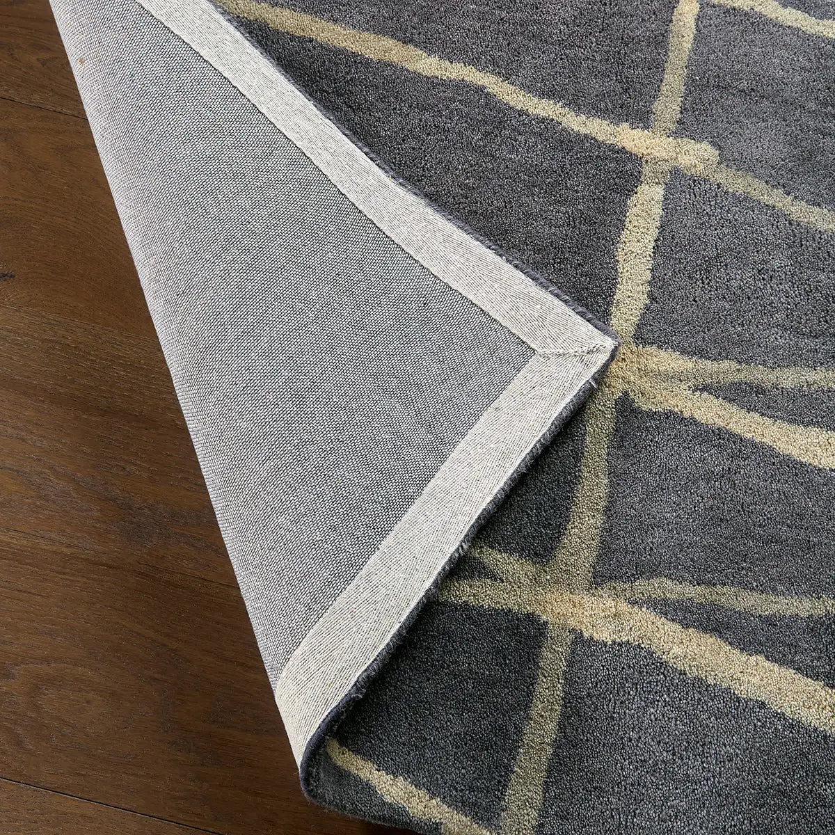 Close-up of a gray area rug with a beige geometric lattice pattern and a light gray binding edge, shown folded over on a dark wood floor.