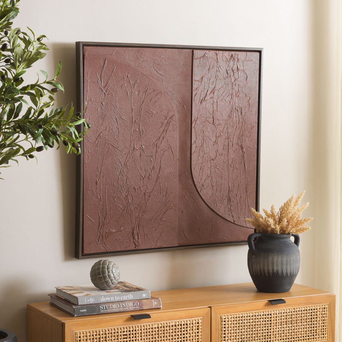 Abstract brown artwork on a wall above a wooden cabinet with books and a vase.